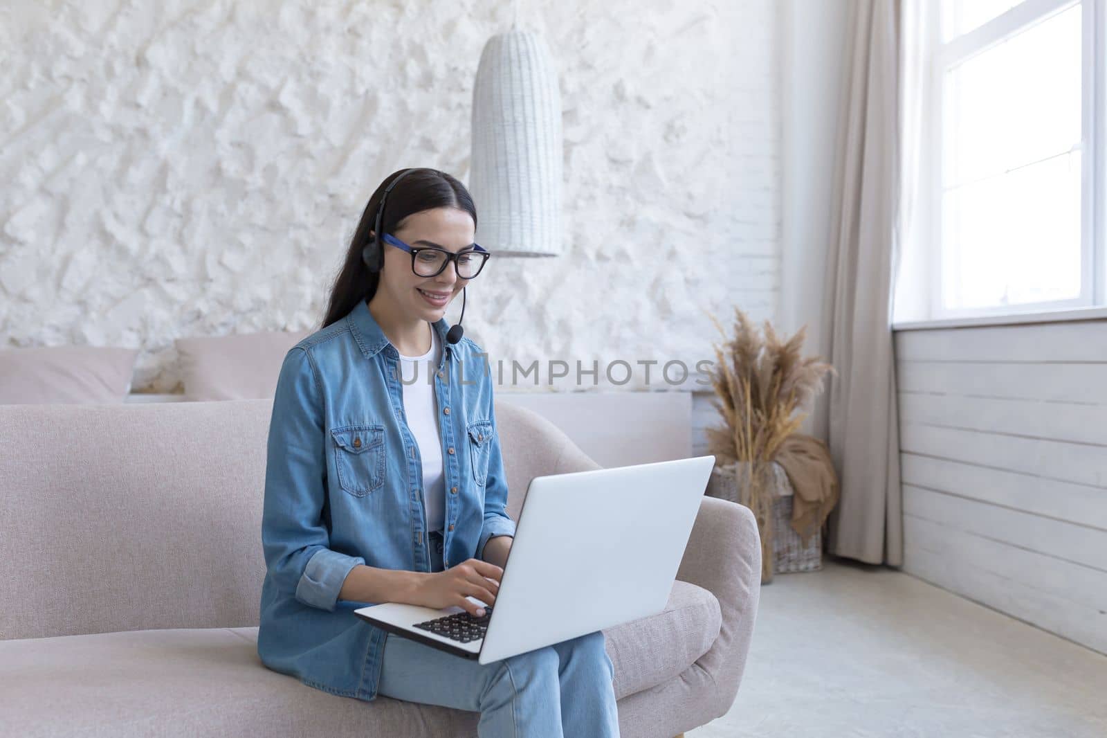 Distance Learning. Portrait of a young smiling female teacher in glasses sitting at home on the sofa in a headset, holding a laptop. Works, teaches from home online.