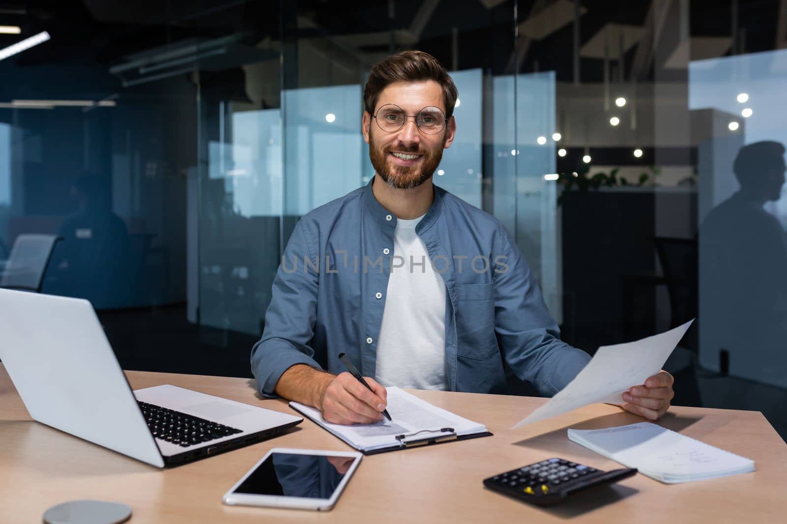 Portrait of a young handsome man, freelancer, businessman, accountant. Sitting in the office, looking at the camera, smiling. Works on a laptop, with documents, fills in forms with a pen.