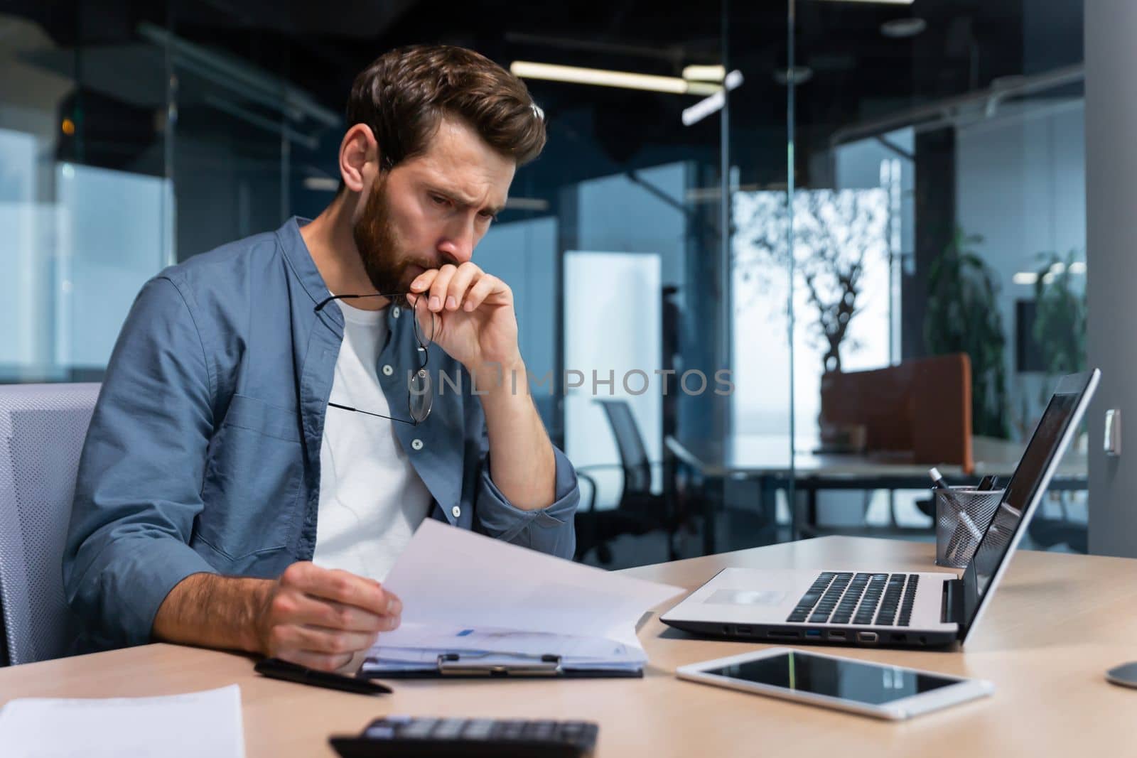 A serious young man accountant, financier, analyst, auditor sits in the office at the table. Thoughtfully holds glasses, looks, analyzes accounts, budget, works with documents and laptop.