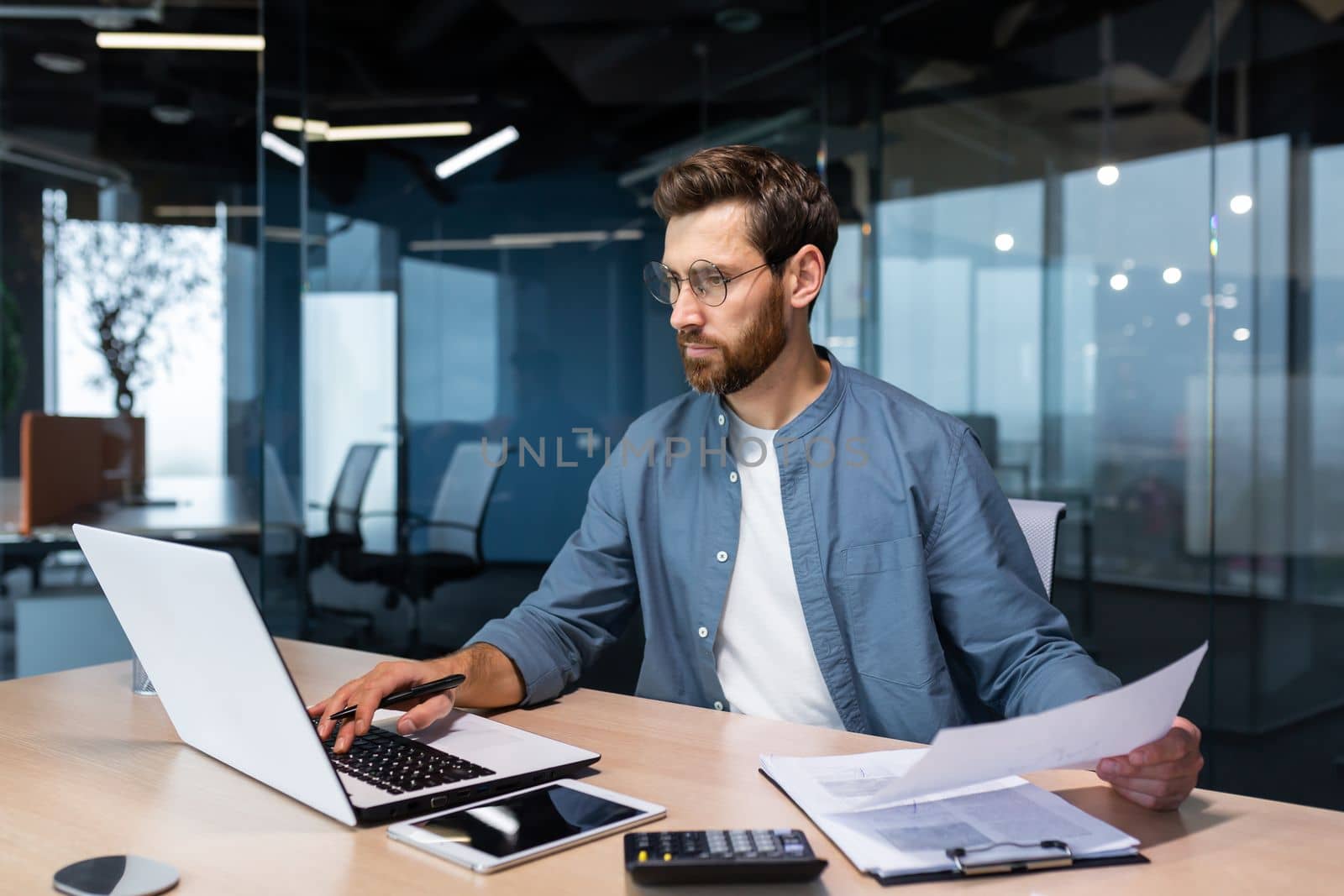 A serious young man accountant, financier, analyst, auditor sits in the office at the table. He holds documents and a pen in his hands, checks accounts, finances, types on a laptop.