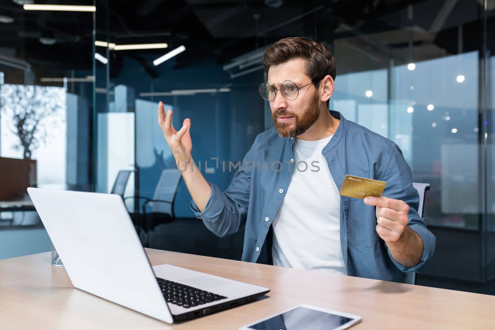 Bankruptcy, financial problems. Angry and worried young man sitting in the office, holding a credit card, looking at a laptop. Checks the account, funds. He waves his arms angrily.