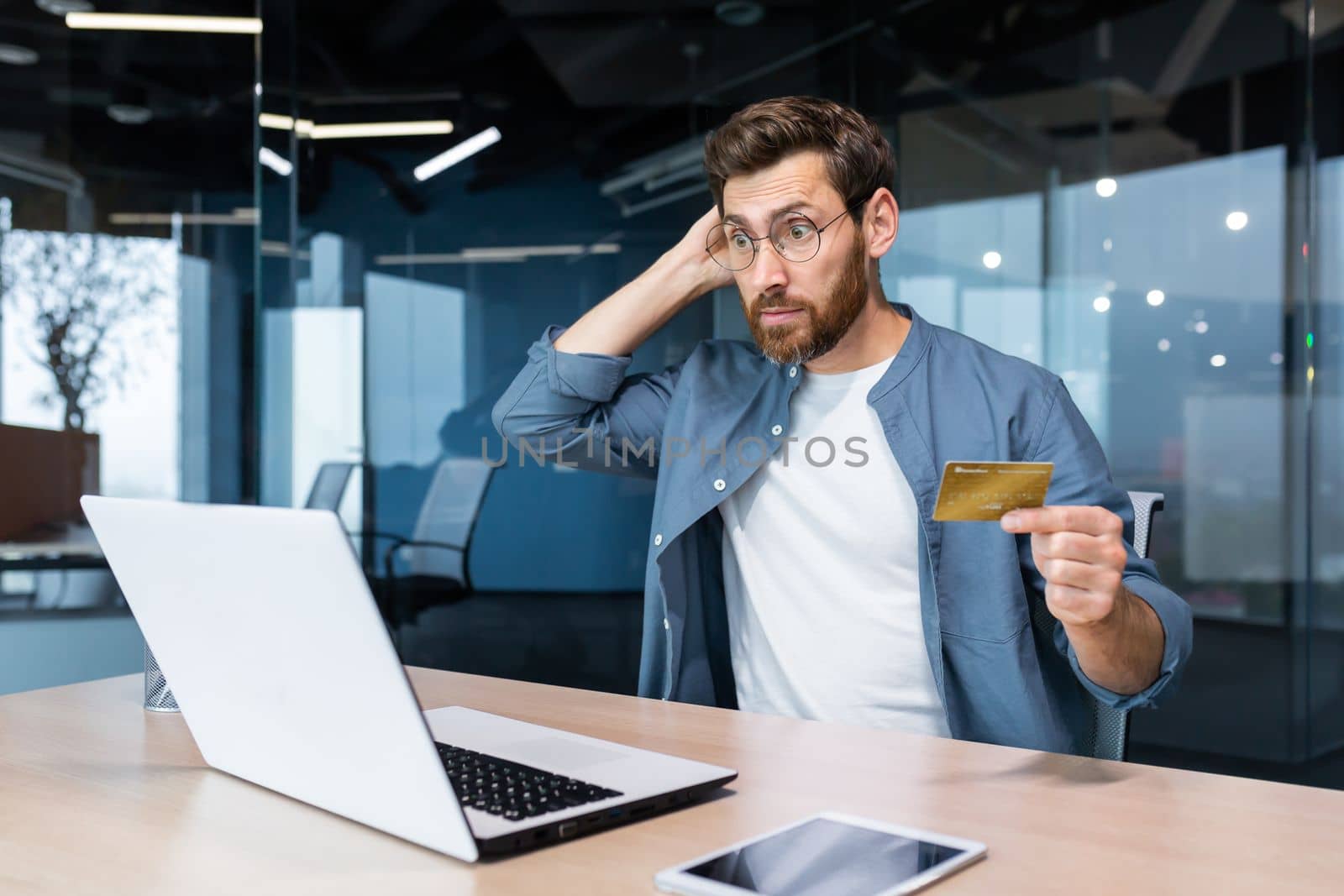 Bankruptcy, financial problems, unsuccessful online shopping. Shocked young man sitting in the office, holding a credit card, looking at a laptop. Checks the account, scratches the back of the head.