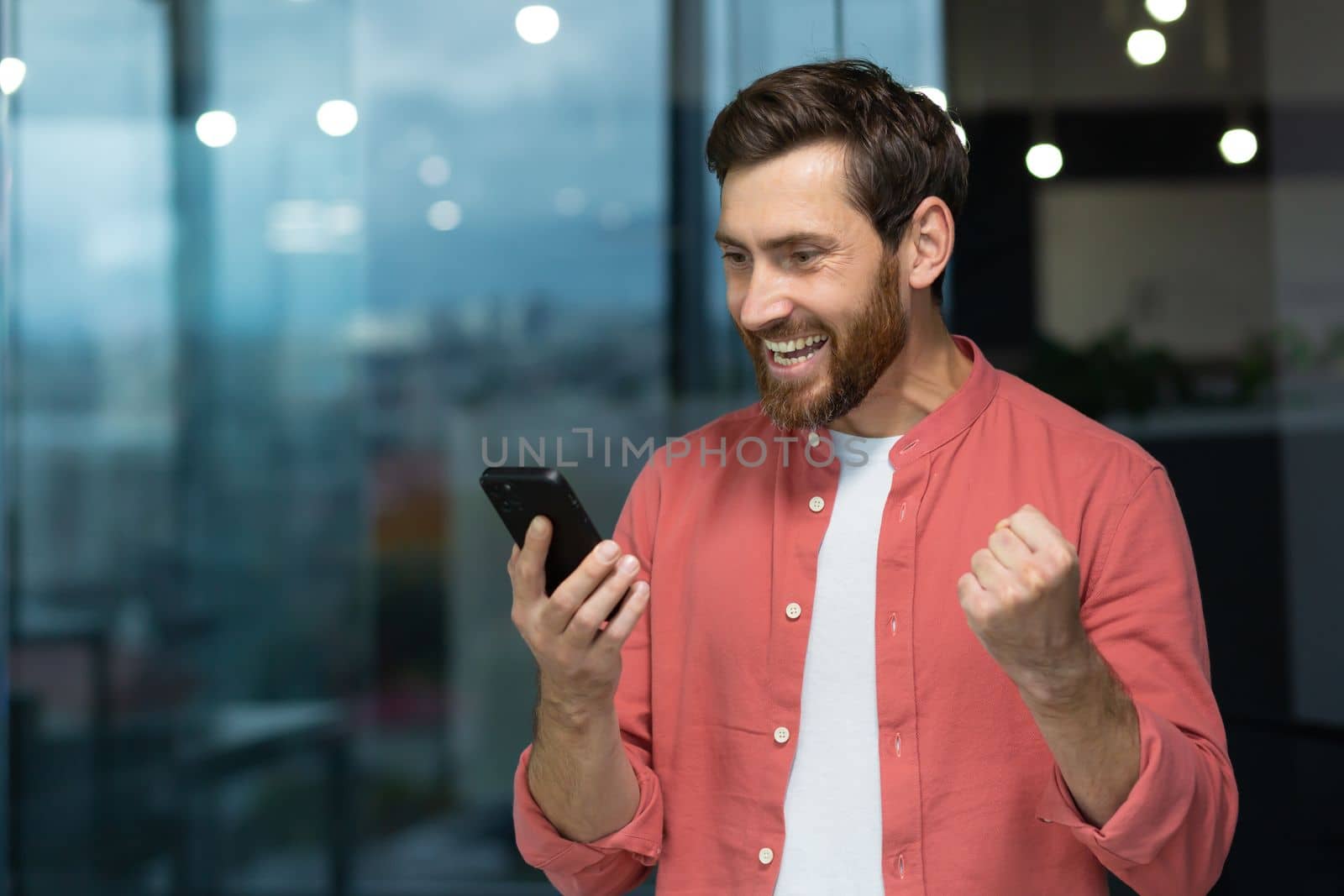 A successful businessman joyfully celebrates victory and good achievements, a man in a red shirt inside the office received an online notification, holding a smartphone in his hands. by voronaman