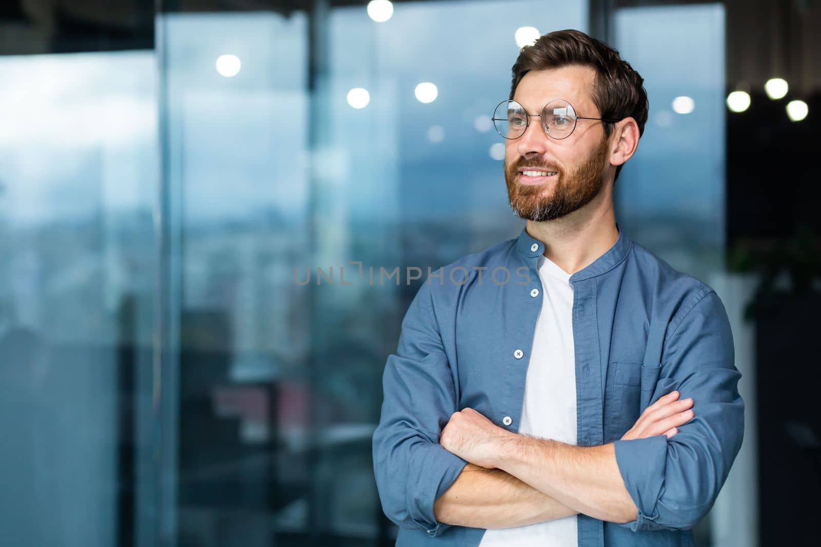 Portrait of a young successful man, businessman, freelancer, designer standing in the office, crossing his arms, confidently looking to the side. by voronaman