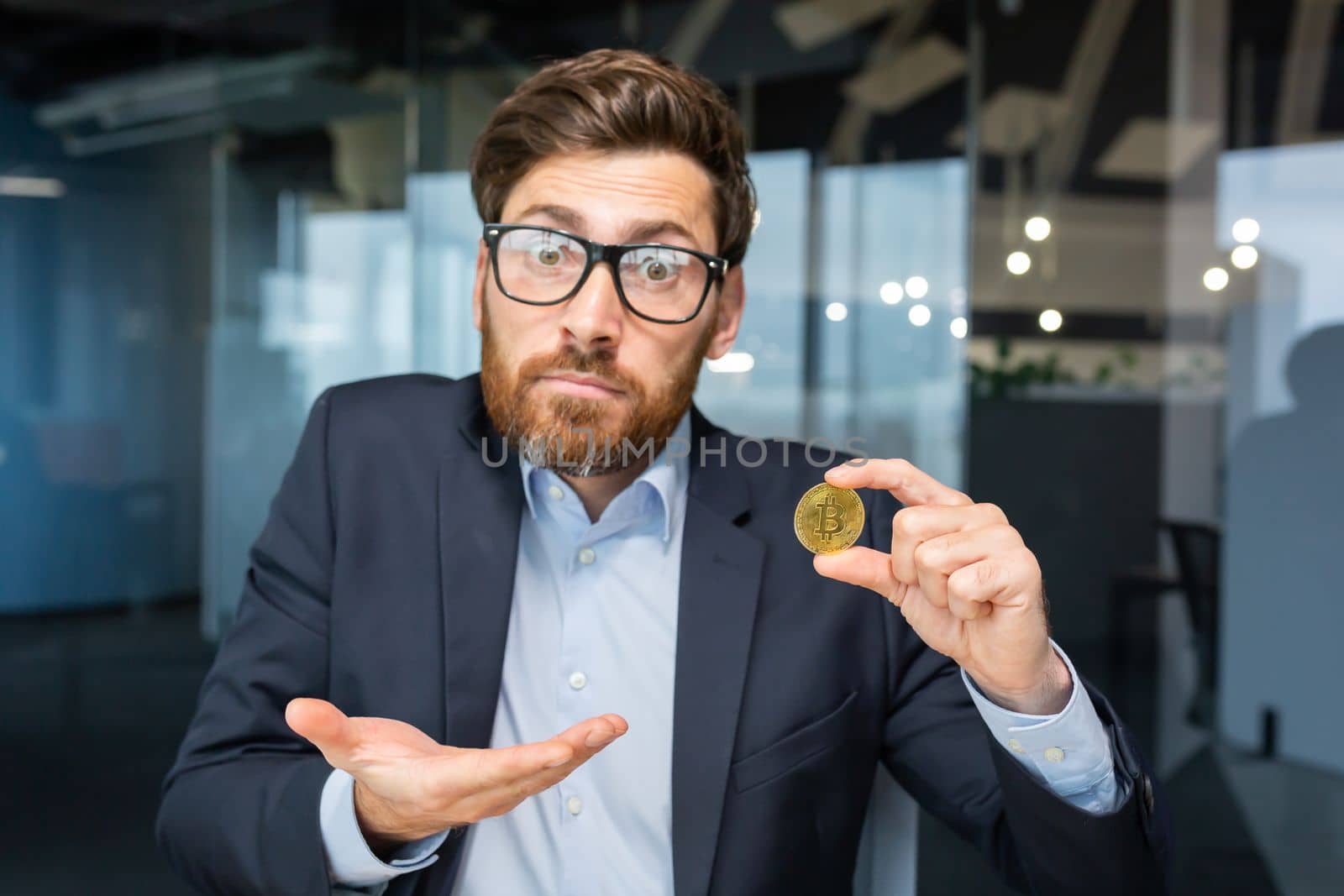 Close-up photo. Confused young business man holds a gold coin, cryptocurrency, bitcoin in his hand. He looks into the camera, points to a coin, points with his hand, asks a question.