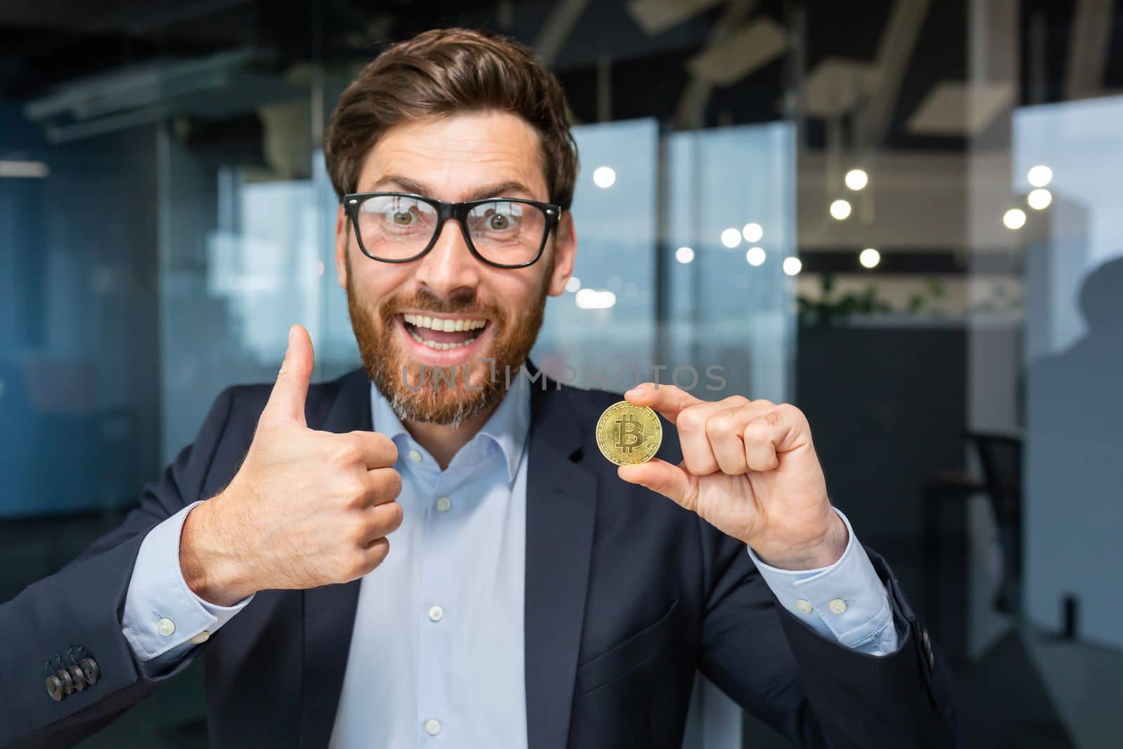 Portrait of mature successful businessman investor, man shows cryptocurrency gold coin to camera and thumbs up firmly, man works on stock exchange inside office online uses laptop.