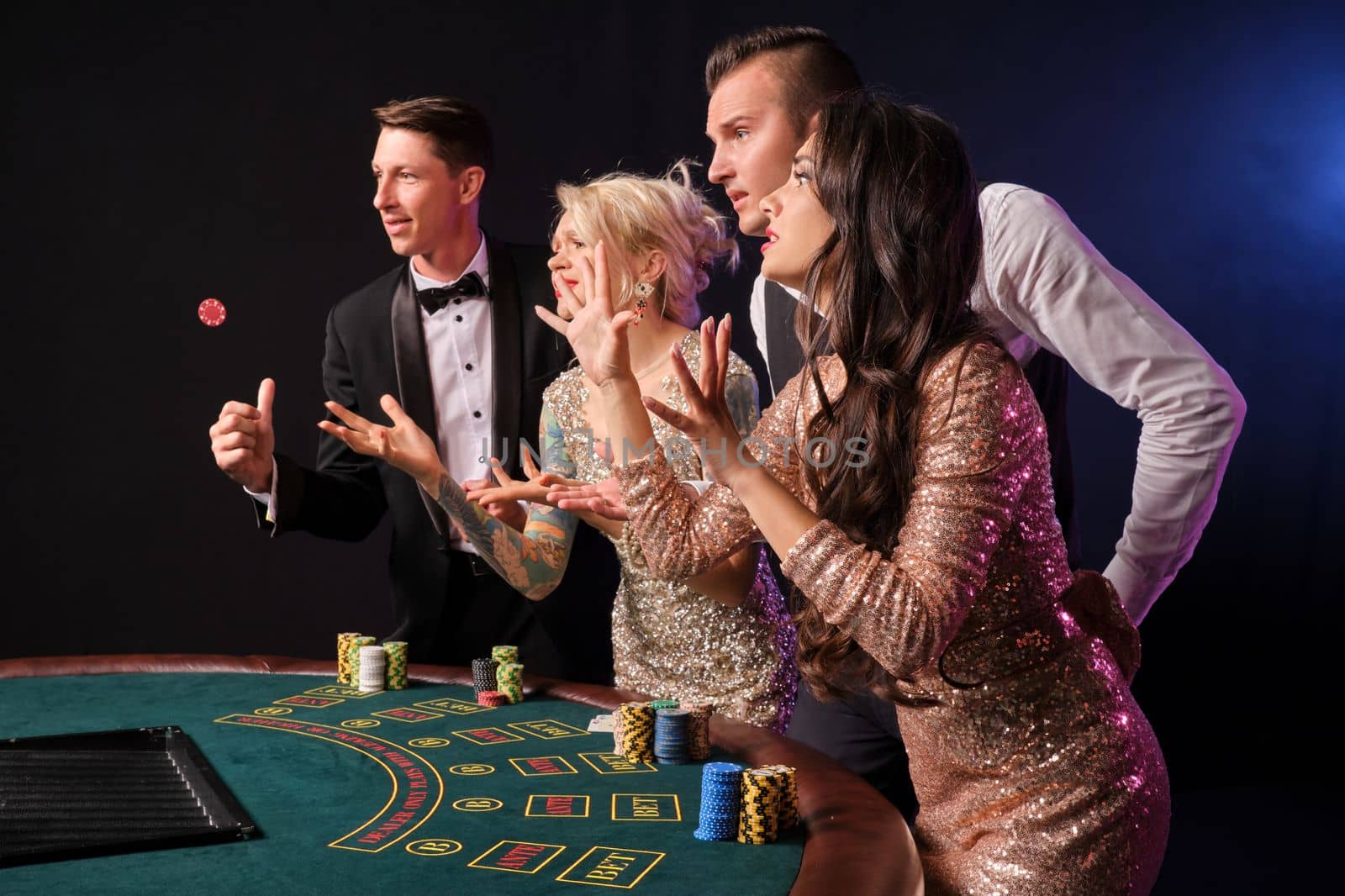 Side shot of a stylish rich friends playing poker at casino. Youth have lose. They are standing at the table against a red and blue backlights on black background. Risky gambling entertainment.