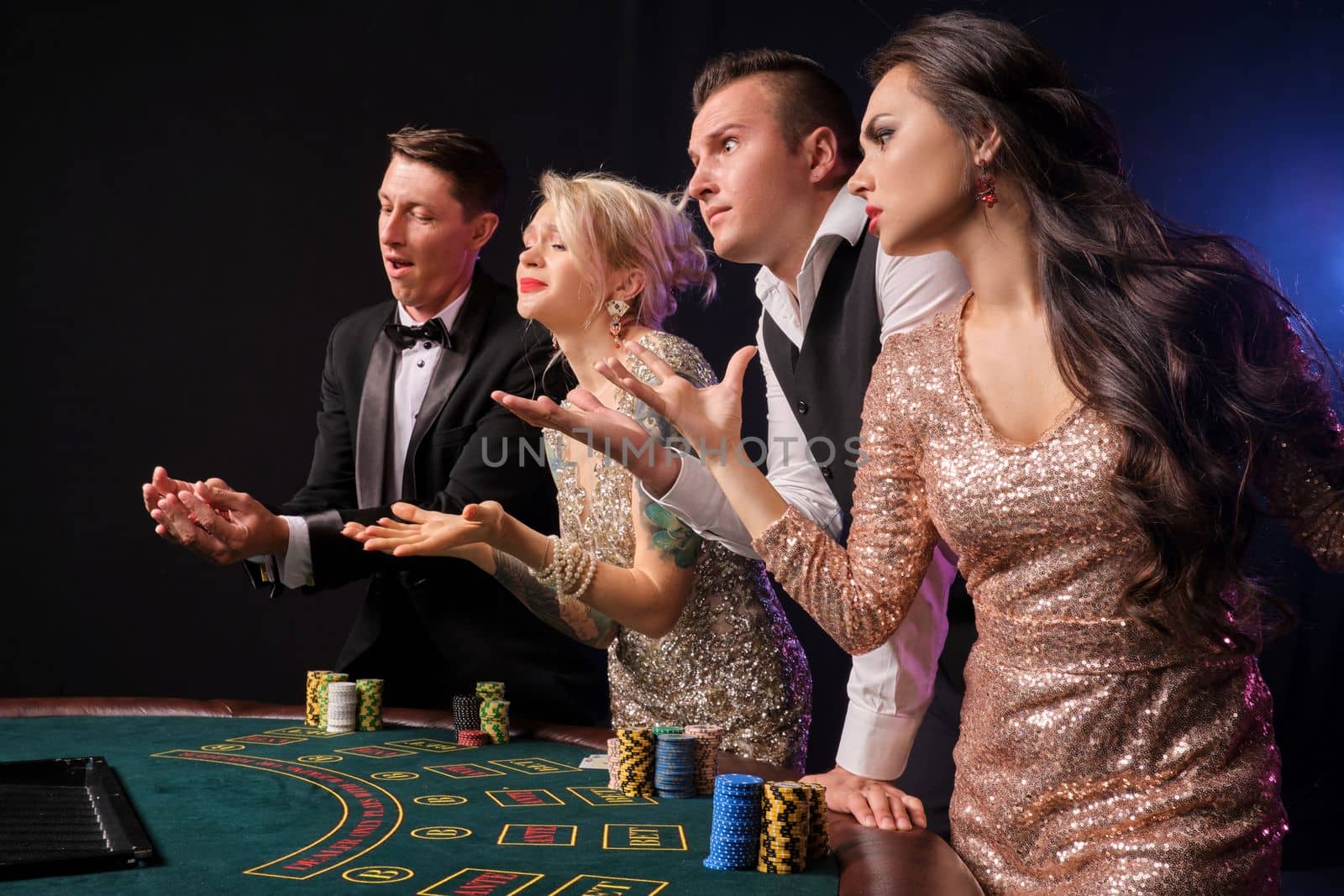 Side shot of a stylish rich friends playing poker at casino. Youth have lose. They are looking angry standing at the table against a red and blue backlights on black background. Risky gambling entertainment.