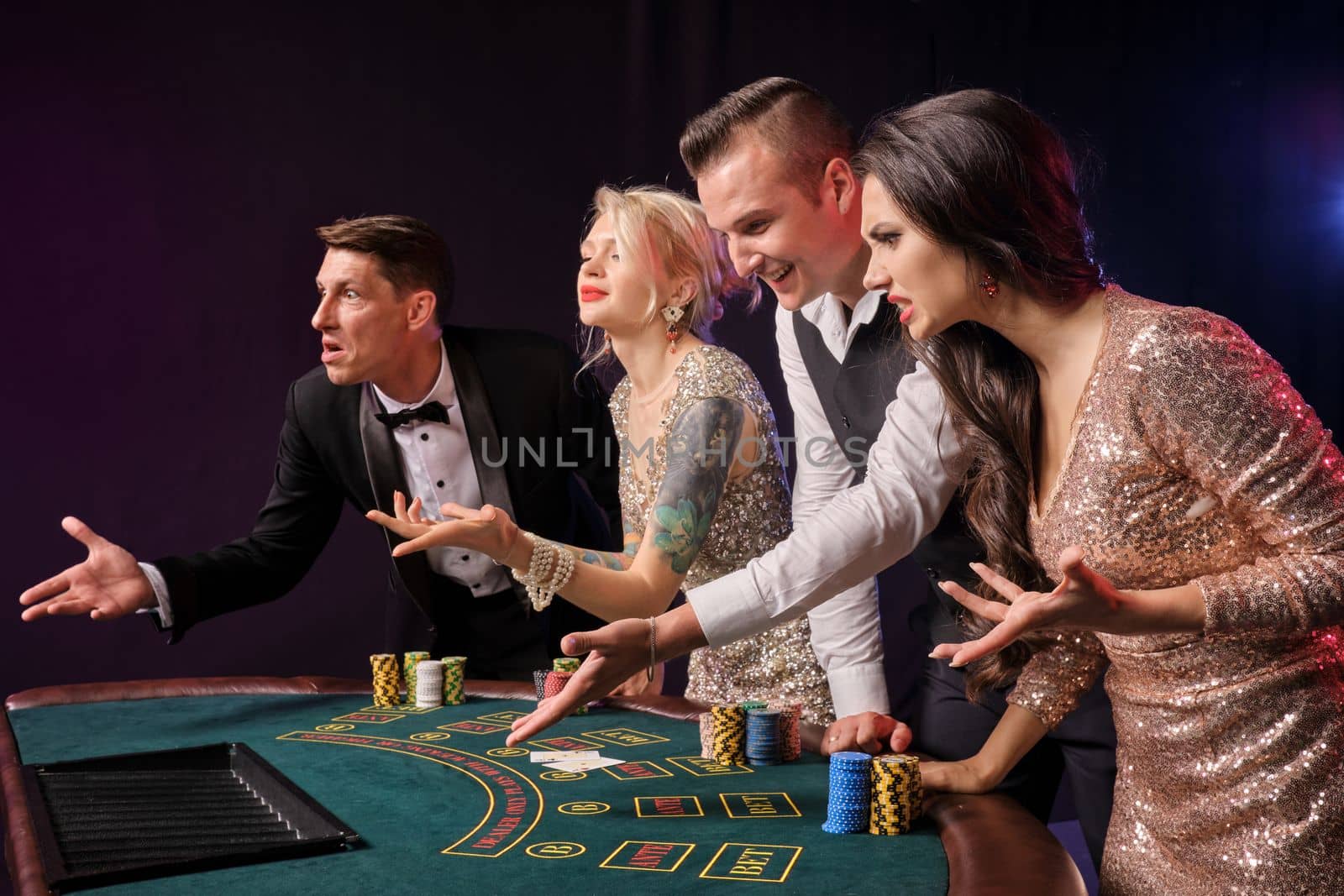 Side shot of a confused rich friends playing poker at casino. Youth have lose. They are looking disappointed standing at the table against a red and blue backlights on black background. Risky gambling entertainment.