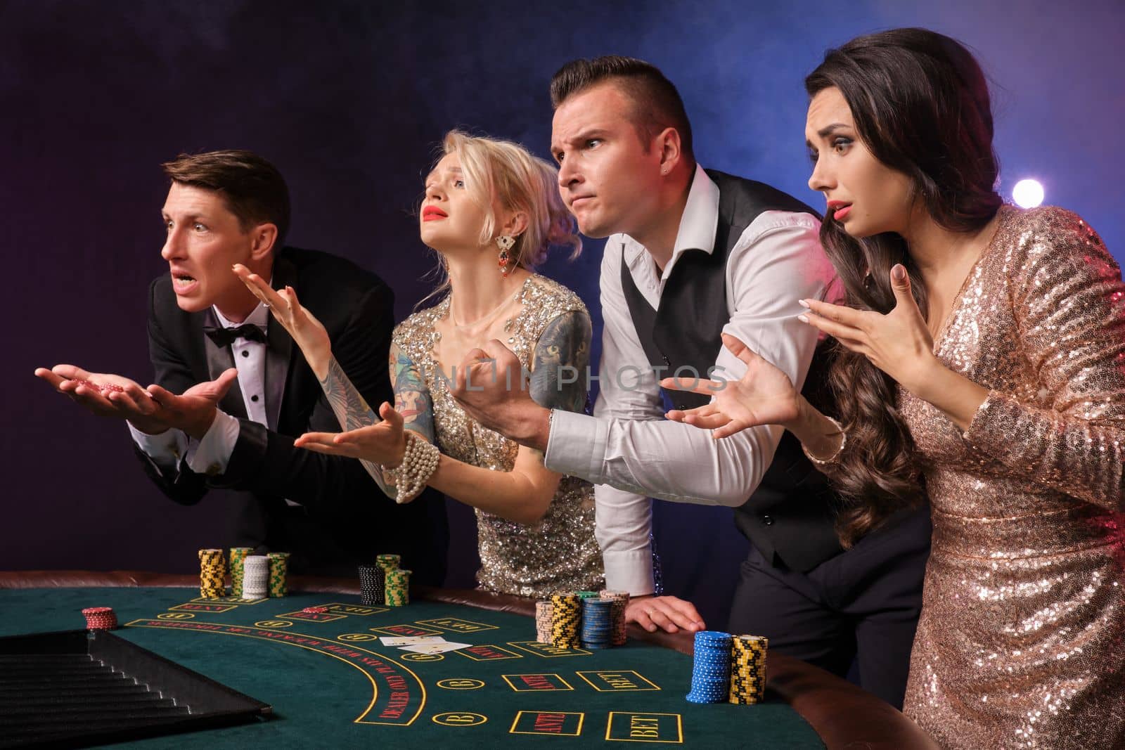 Side shot of an upset rich companions playing poker at casino in smoke. Youth are making bets waiting for a big win. They are standing at the table against a red and blue backlights on black background. Risky gambling entertainment.