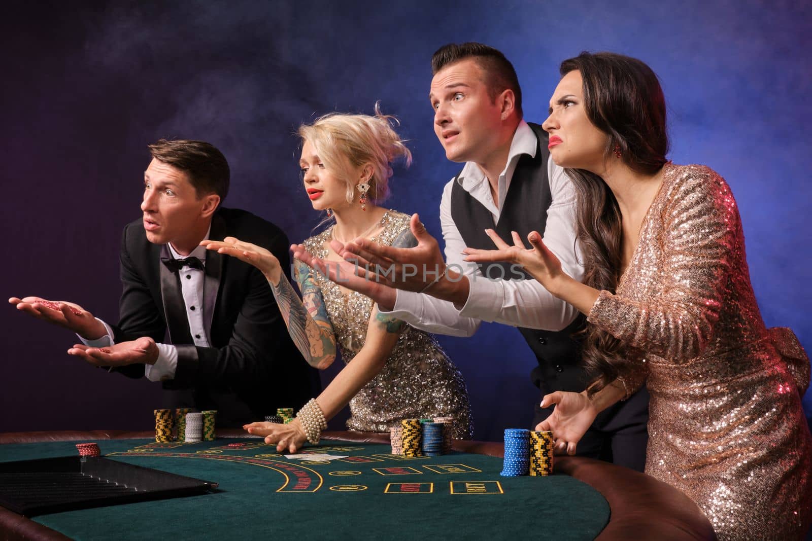 Side shot of an upset rich colleagues playing poker at casino in smoke. Youth are making bets waiting for a big win. They are looking unhappy standing at the table against a red and blue backlights on black background. Risky gambling entertainment.