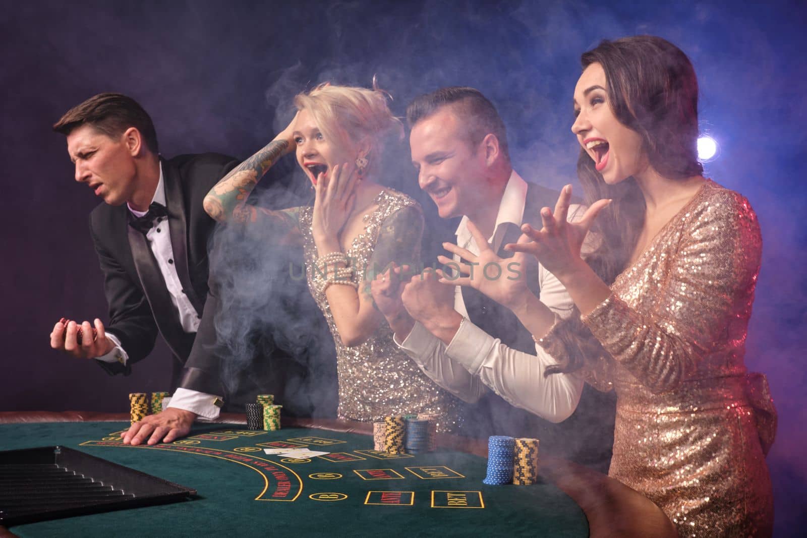 Side shot of an overjoyed rich companions playing poker at casino in smoke. Youth are making bets waiting for a big win. They are celebrating their victory standing at the table against a red and blue backlights on black background. Risky gambling entertainment.