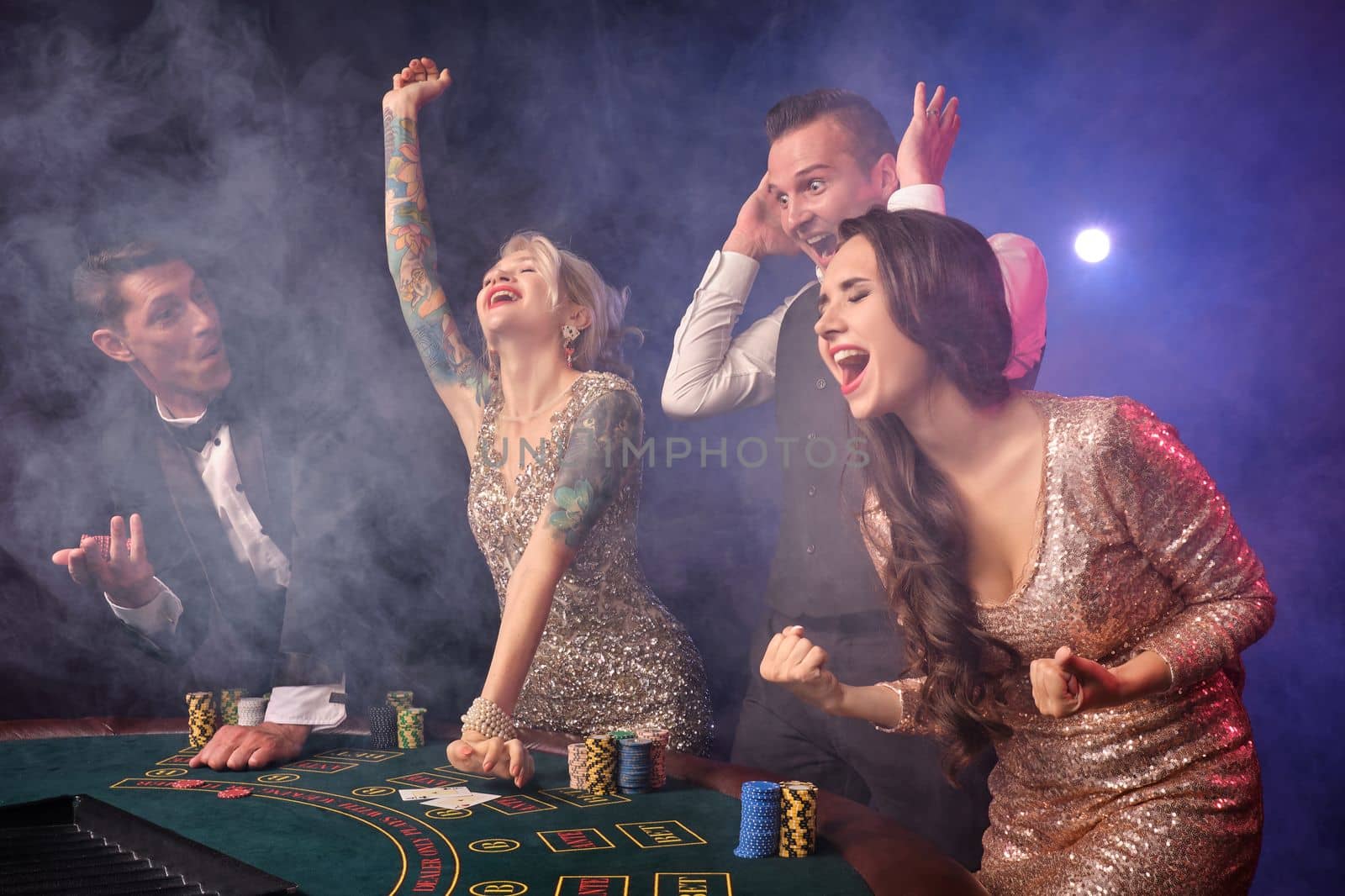 Side shot of an overjoyed rich colleagues playing poker at casino in smoke. Youth are making bets waiting for a big win. They are looking overjoyed standing at the table against a red and blue backlights on black background. Risky gambling entertainment.