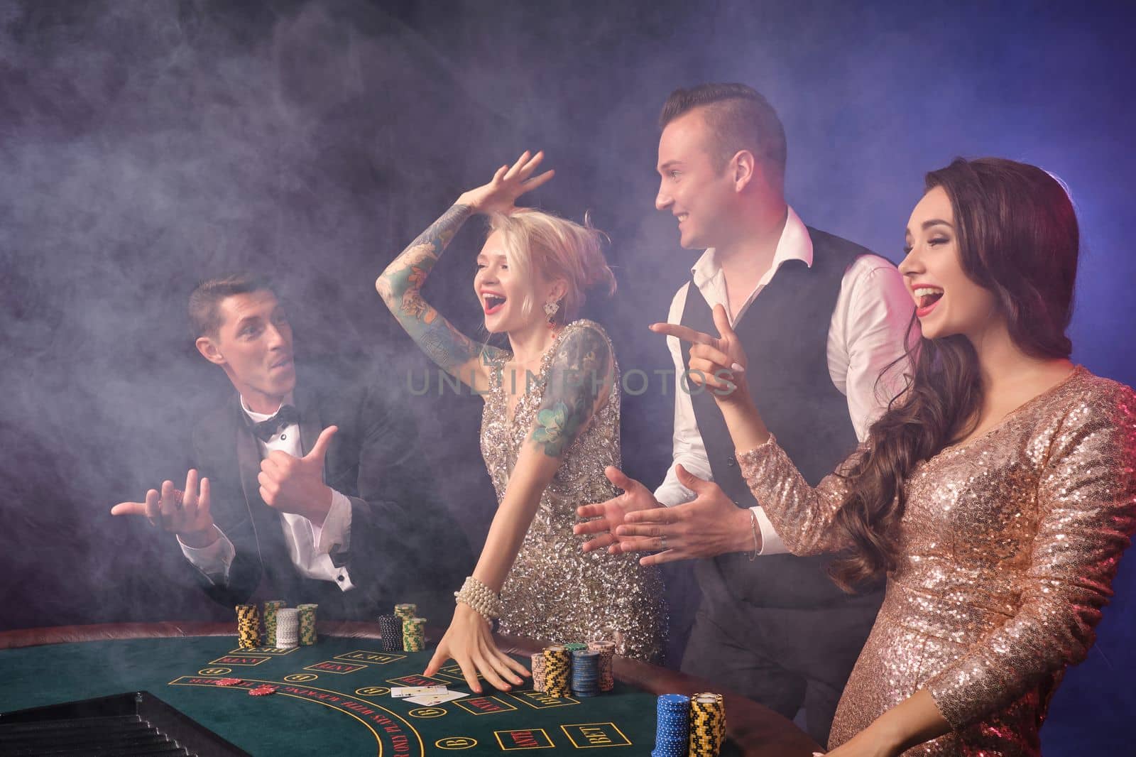 Side shot of an overjoyed rich classmates playing poker at casino in smoke. Youth are making bets waiting for a big win. They are smiling standing at the table against a red and blue backlights on black background. Risky gambling entertainment.
