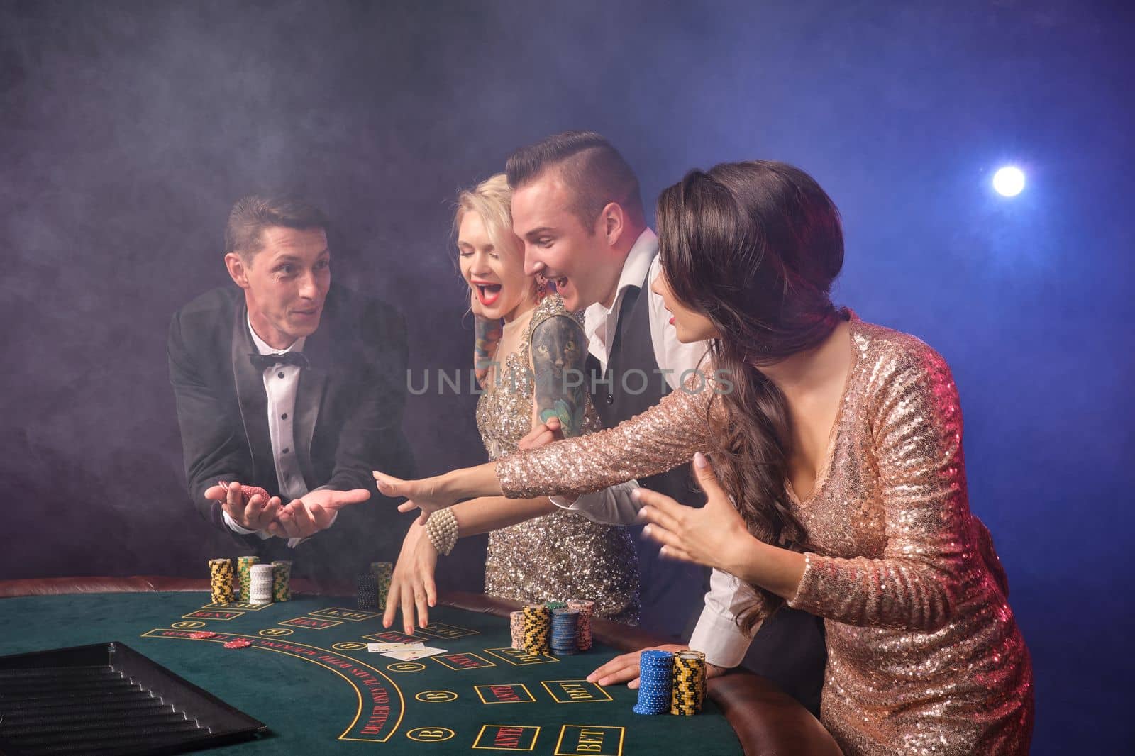 Side shot of a joyful rich buddies playing poker at casino in smoke. Youth are making bets waiting for a big win. They are standing at the table against a red and blue backlights on black background. Risky gambling entertainment.