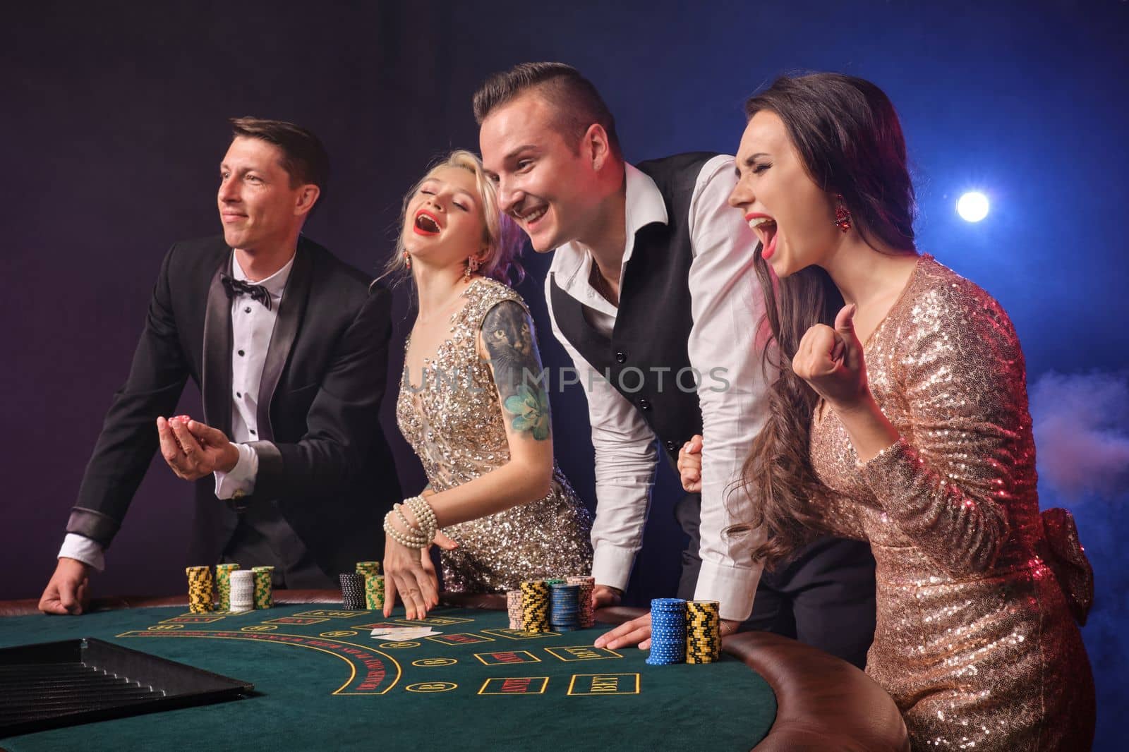 Side shot of a joyful rich companions playing poker at casino in smoke. Youth are making bets waiting for a big win. They are looking excited standing at the table against a red and blue backlights on black background. Risky gambling entertainment.