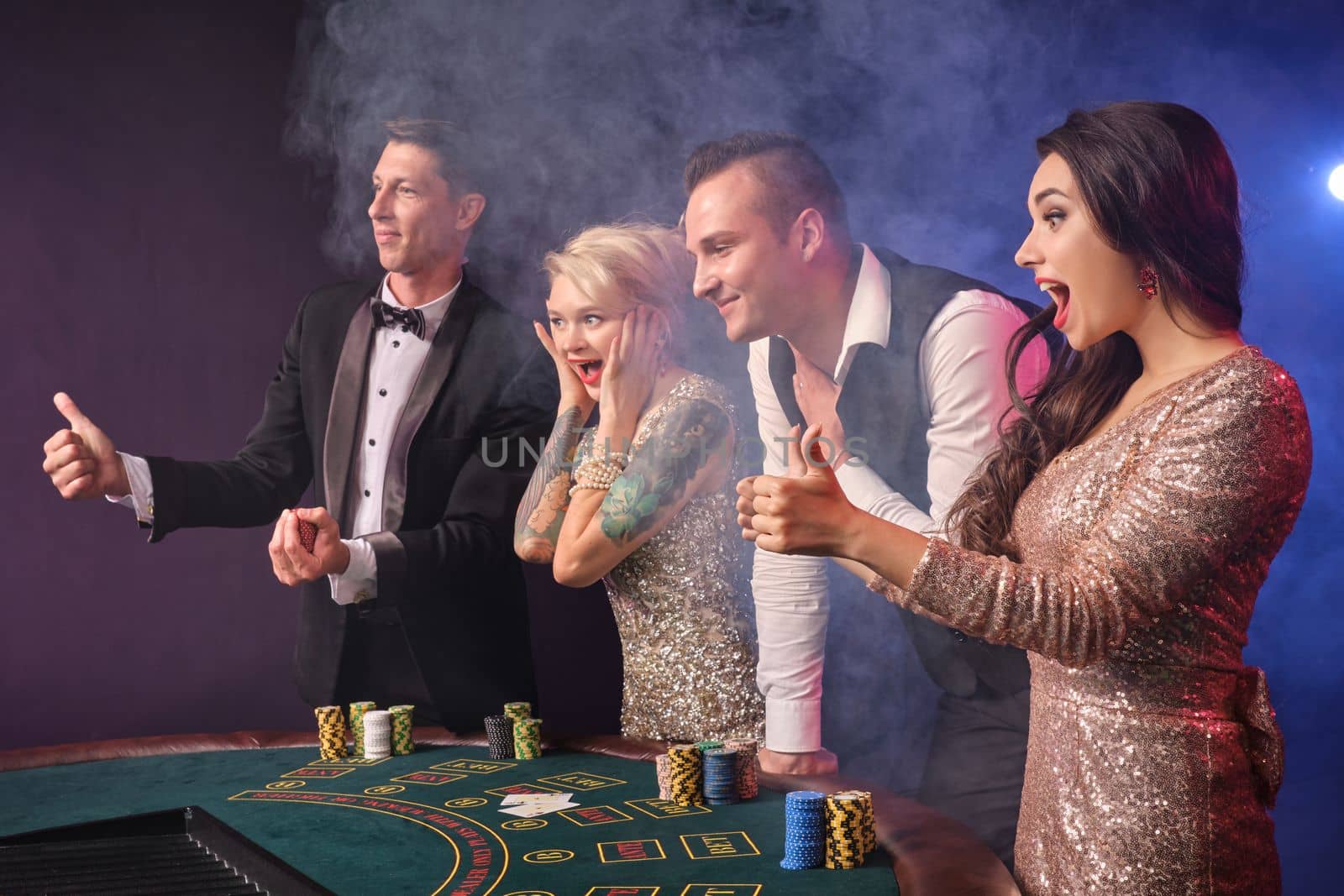 Side shot of a joyful rich colleagues playing poker at casino in smoke. Youth are making bets waiting for a big win. They are looking happy standing at the table against a red and blue backlights on black background. Risky gambling entertainment.