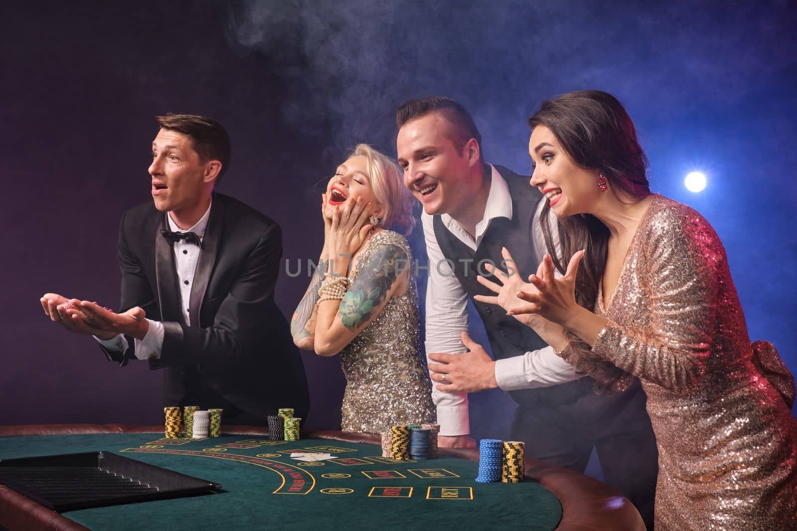 Side shot of a joyful rich classmates playing poker at casino in smoke. Youth are making bets waiting for a big win. They are laughing standing at the table against a red and blue backlights on black background. Risky gambling entertainment.