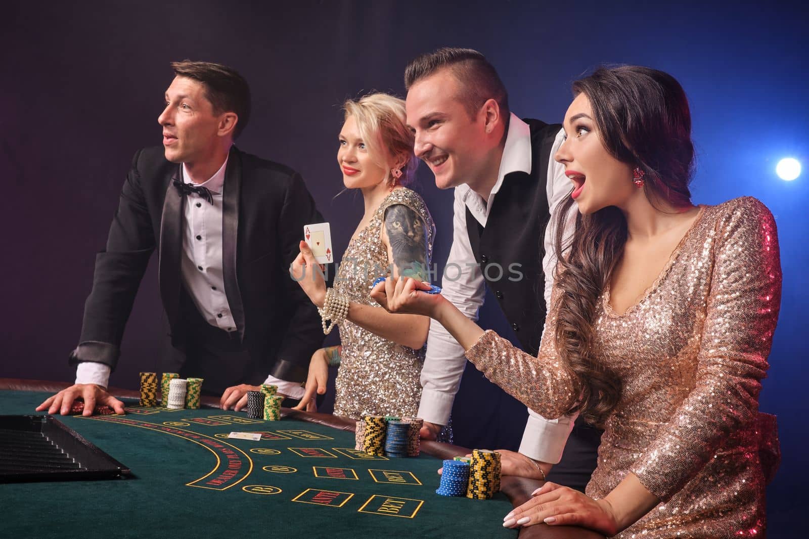Side shot of a cheerful rich companions playing poker at casino. Youth are making bets waiting for a big win. They are looking away standing at the table against a red and blue backlights on black background. Risky gambling entertainment.
