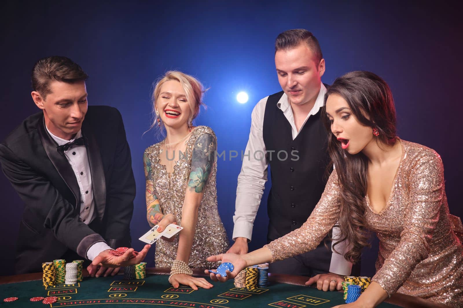 Group of a cheerful rich colleagues are playing poker at casino. Youth are making bets waiting for a big win. They are standing at the table against a red and blue backlights on black background. Risky gambling entertainment.