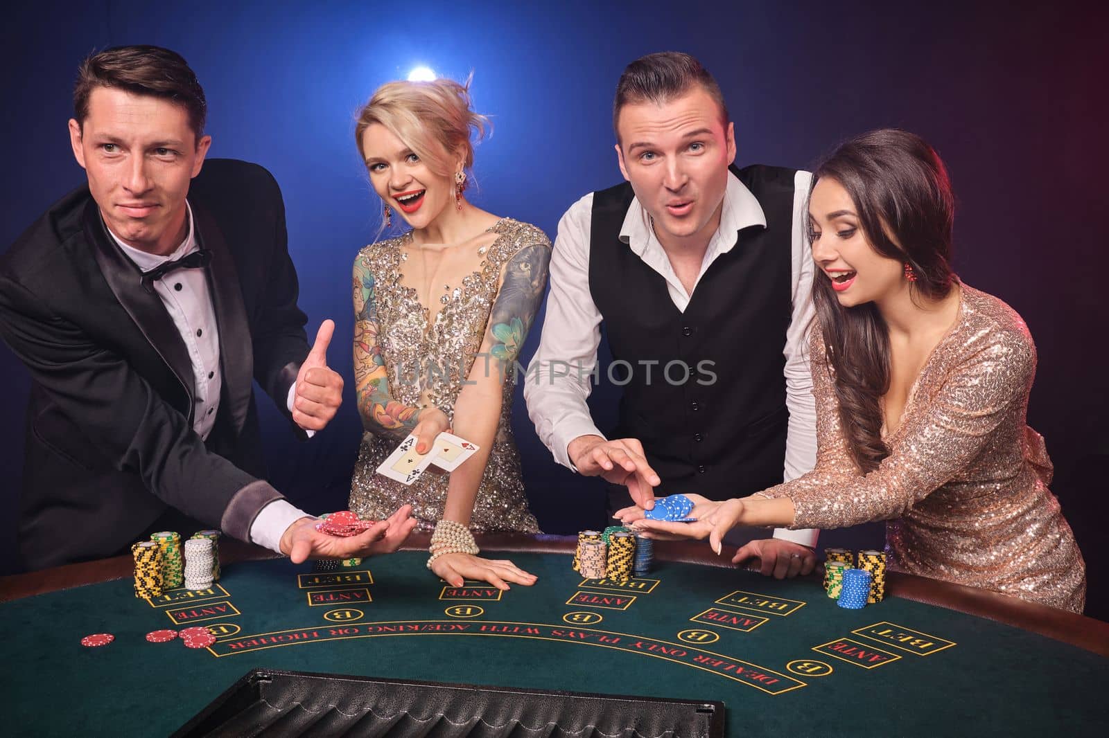 Group of a cheerful rich friends are playing poker at casino. Youth are making bets waiting for a big win. They are standing at the table against a red and blue backlights on black background. Risky gambling entertainment.