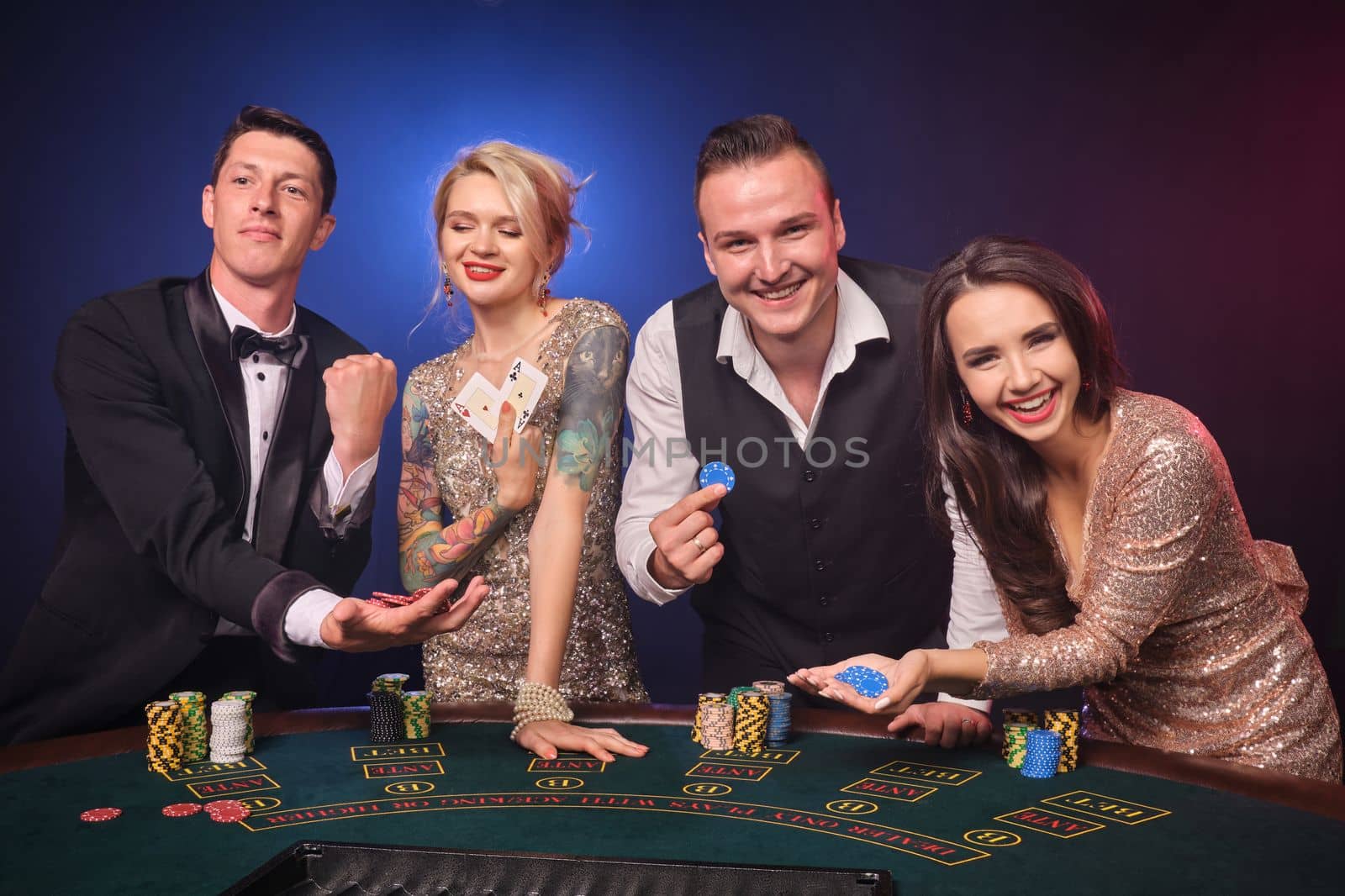 Group of an excited rich buddies are playing poker at casino. Youth are making bets waiting for a big win. They are smiling standing at the table against a red and blue backlights on black background. Risky gambling entertainment.
