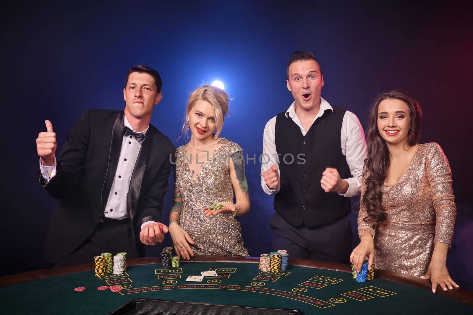 Group of an excited rich colleagues are playing poker at casino. Youth are making bets waiting for a big win. They are looking happy standing at the table against a red and blue backlights on black background. Risky gambling entertainment.