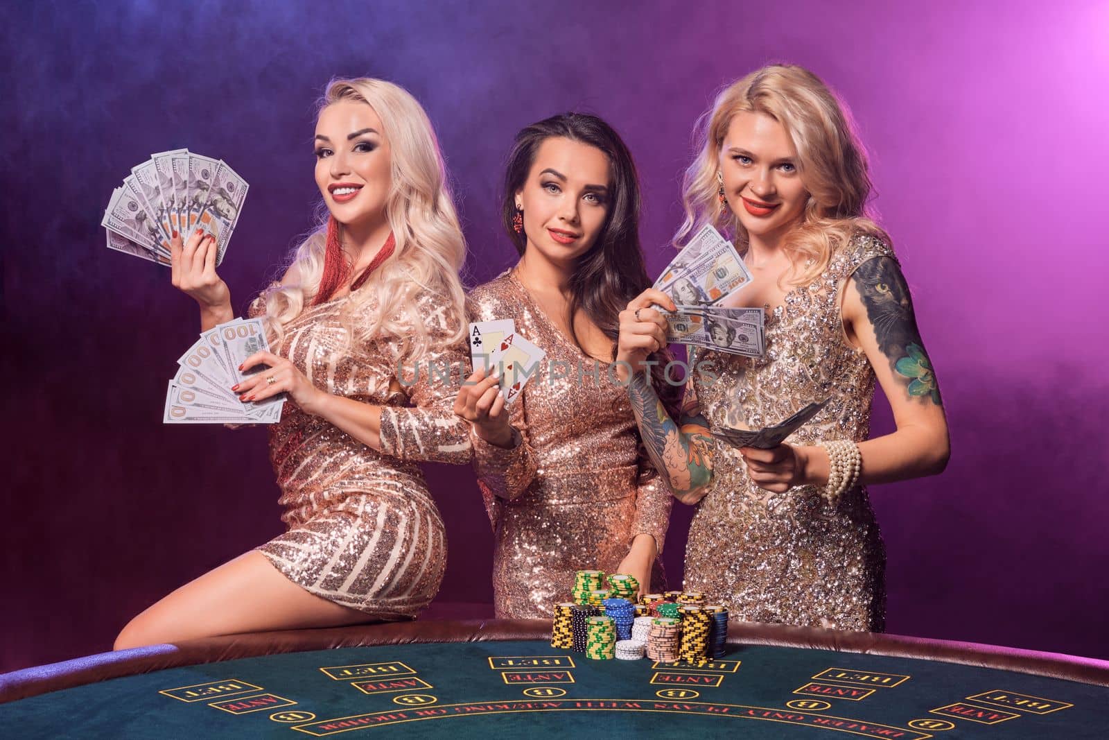 Beautiful sisters with a perfect hairstyles and bright make-up, dressed in a golden shiny dresses are posing standing at a gambling table and smiling. Poker concept on a black smoke background with pink and blue backlights. Casino.