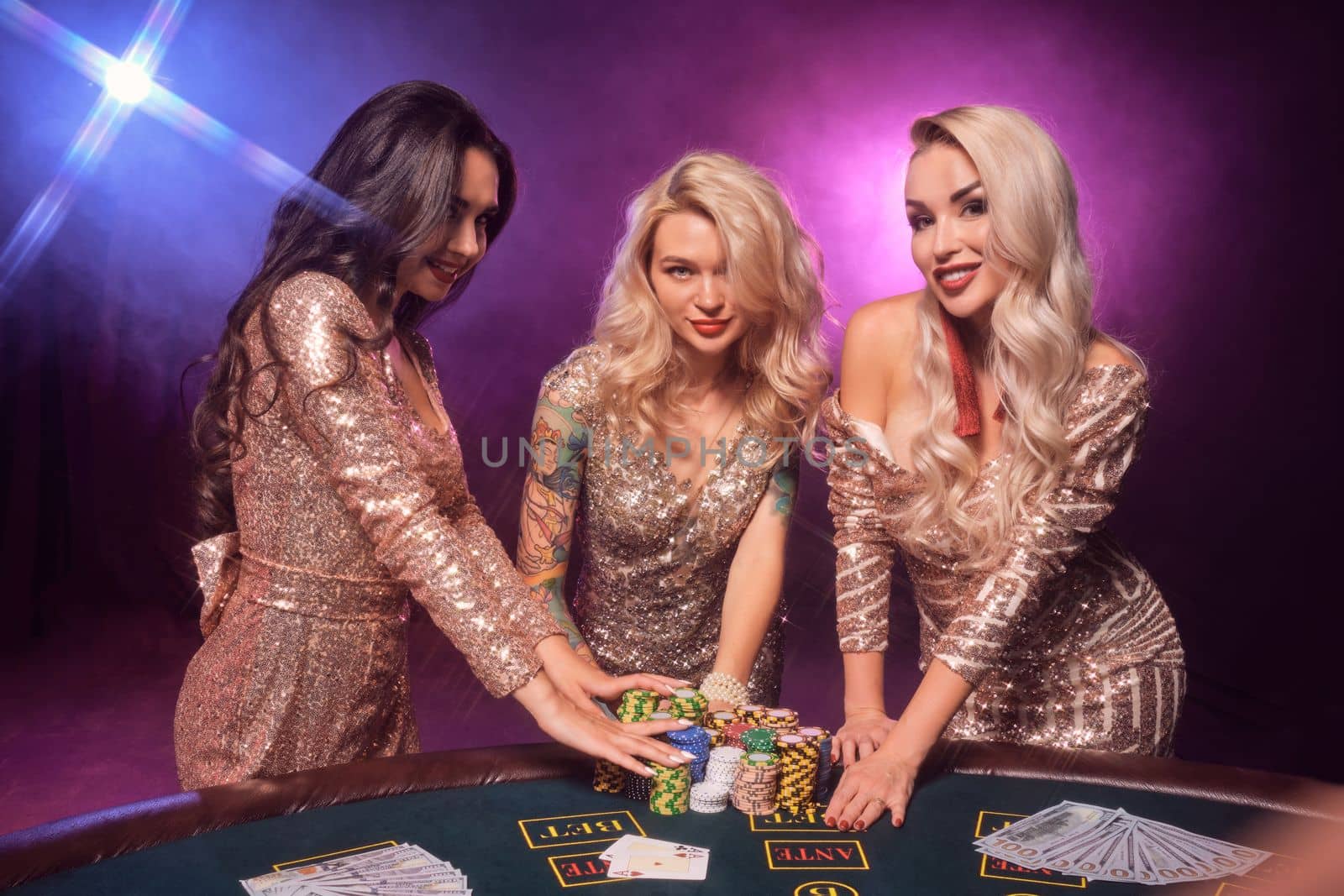 Beautiful girls with a perfect hairstyles and bright make-up, dressed in a golden shiny dresses are posing standing at a gambling table and looking very hot. Poker concept on a black smoke background with pink and blue backlights. Casino.