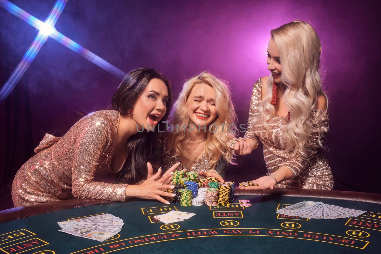 Beautiful females with a perfect hairstyles and bright make-up, dressed in a golden shiny dresses are posing standing at a gambling table and laughing. Poker concept on a black smoke background with pink and blue backlights. Casino.