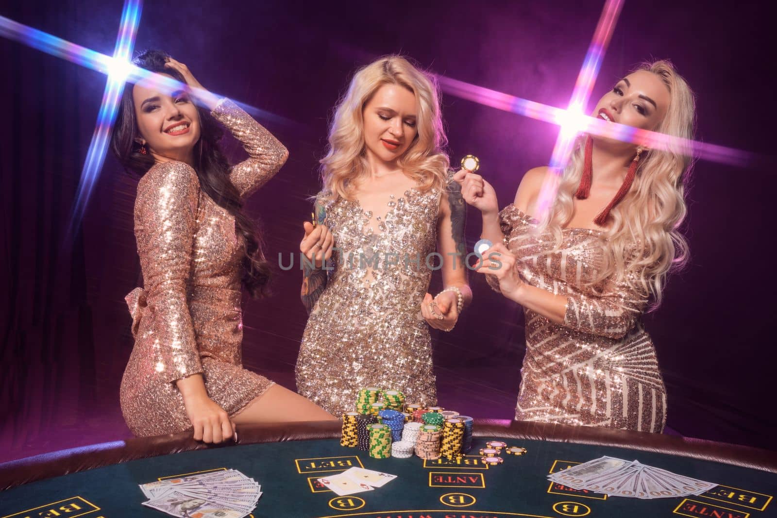 Beautiful women with a perfect hairstyles and bright make-up, dressed in a golden shiny dresses are posing standing at a gambling table. Poker concept on a black smoke background with pink and blue backlights. Casino.