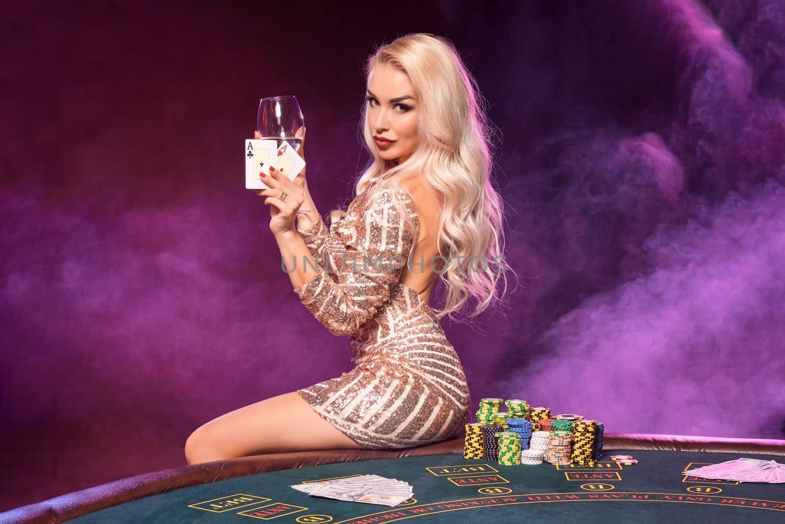 Close-up shot of a charming maiden with blond hair and bright make-up, dressed in a golden striped shiny dress, posing with two aces and glass of champagne in her hands while sitting on a table with chips on it. Poker concept on a black smoke background with pink and blue backlights. Casino.