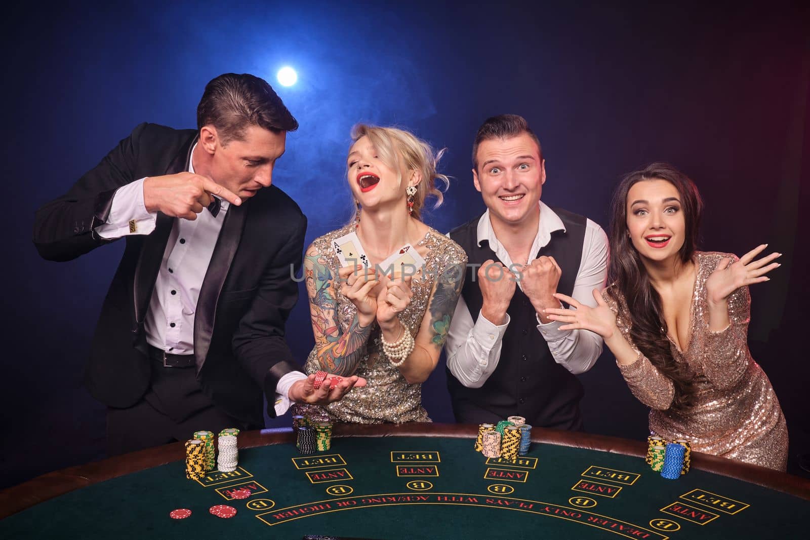 Group of a happy rich buddies are playing poker at casino. Youth are making bets waiting for a big win. They are looking happy standing at the table against a red and blue backlights on black background. Risky gambling entertainment.