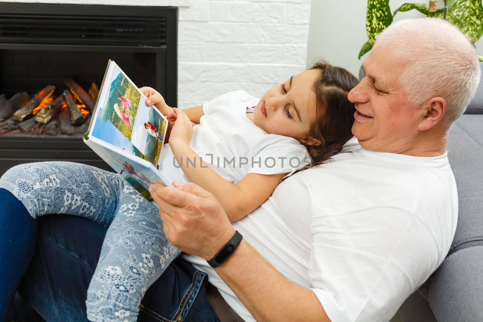 Grandfather And Granddaughter Looking Through Photo Album In Lounge At Home Together by Andelov13