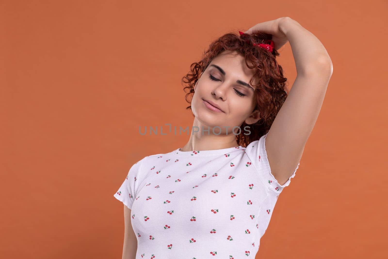 Curly chestnut-colored hair. A woman corrects her hair gently with her hand. White blouse with a cherry pattern. Isolated from the uniform background.