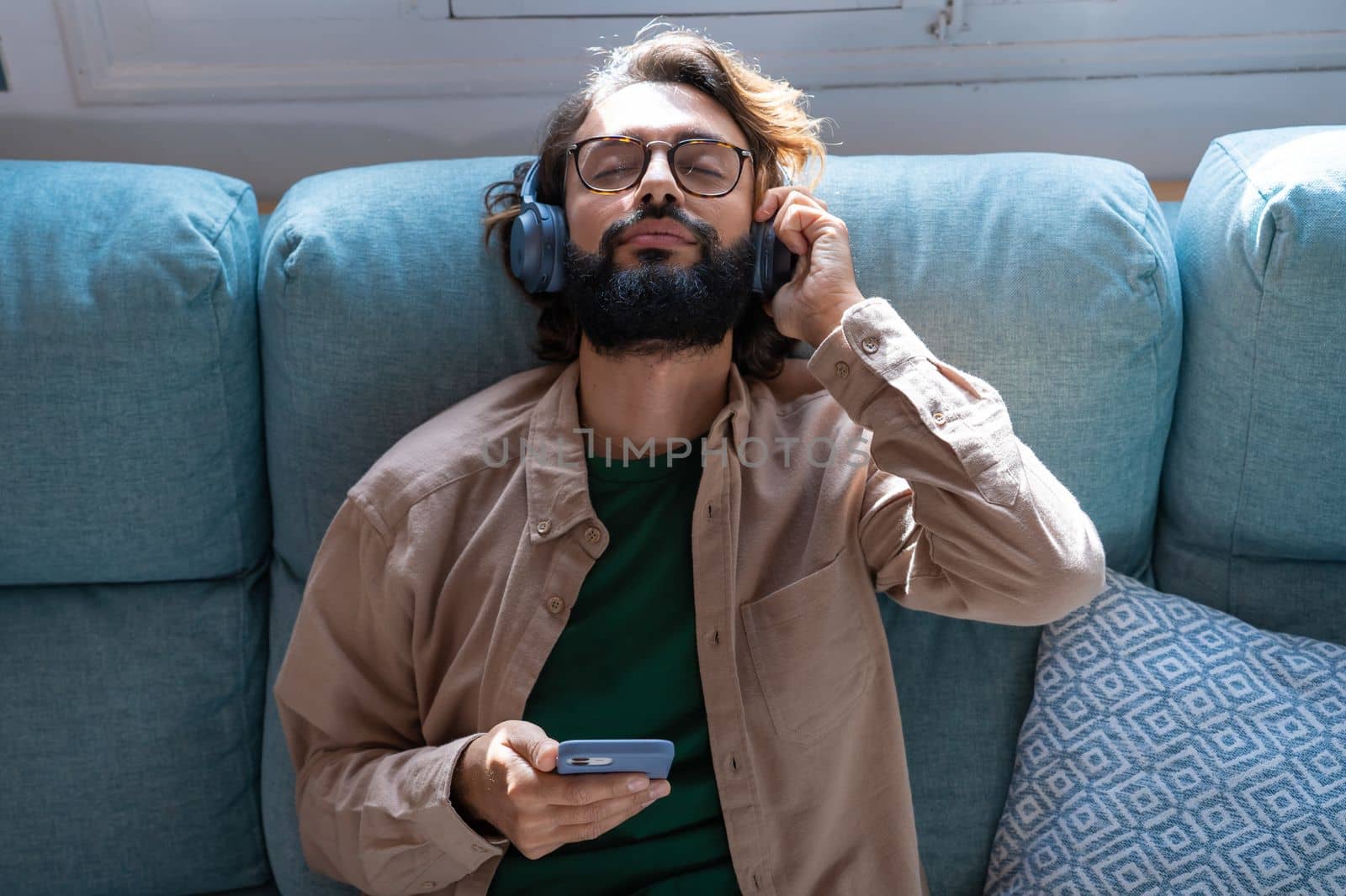 Young man wearing headphones listen podcast or music playing in smartphone sitting on sofa at home by PaulCarr