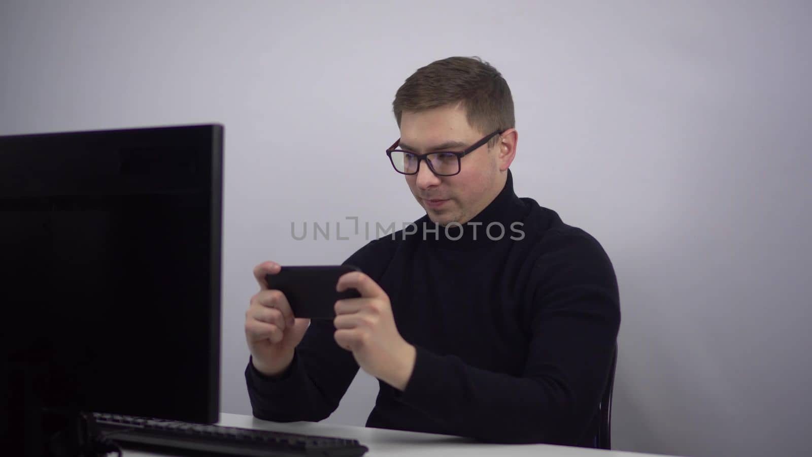 The young man is enthusiastically playing his smartphone and emotionally rejoices at the victory. IT specialist in glasses with a phone in his hands at the workplace in the office. 4k