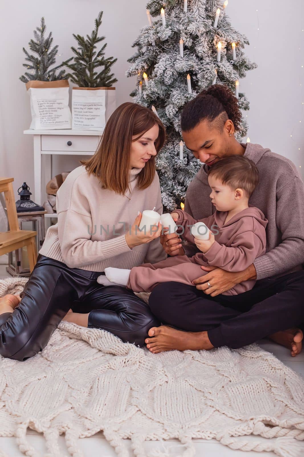 A young diverse family with a baby boy is sitting on a knitted blanket near a Christmas tree by Zakharova