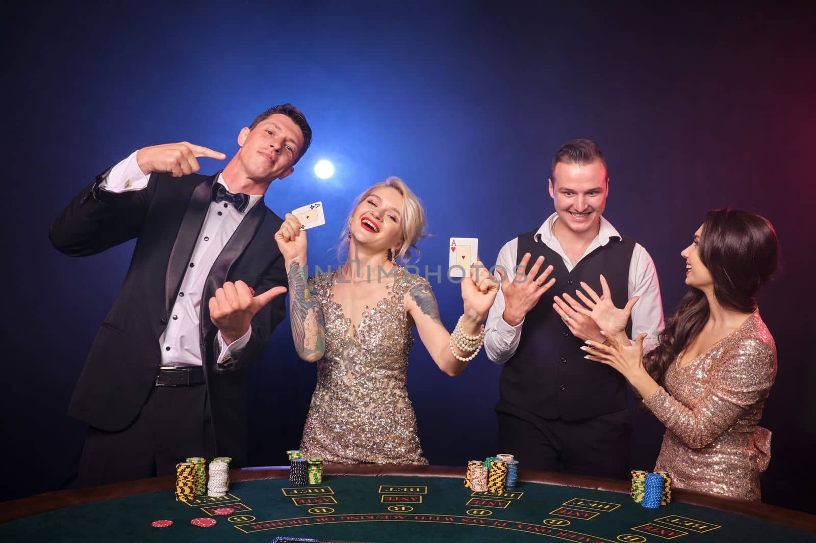 Group of an elegant rich buddies are playing poker at casino. Youth are making bets waiting for a big win. They are looking excited standing at the table against a red and blue backlights on black background. Risky gambling entertainment.