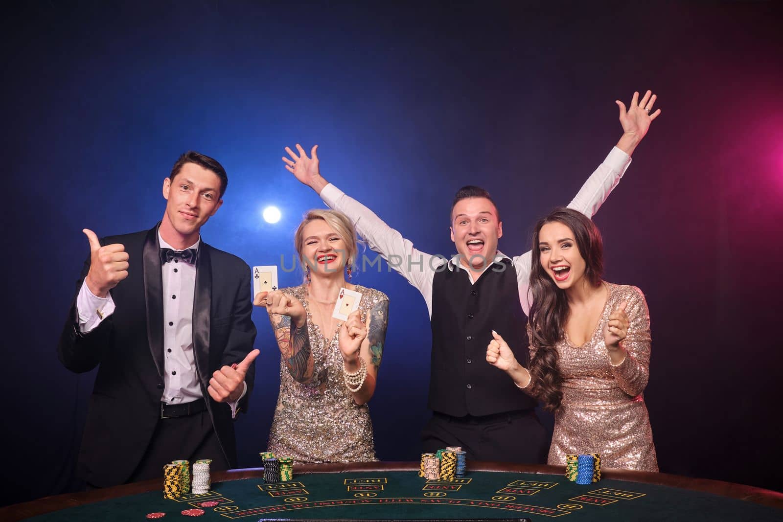 Group of an elegant rich colleagues are playing poker at casino. Youth are making bets waiting for a big win. They are smiling standing at the table against a red and blue backlights on black background. Risky gambling entertainment.