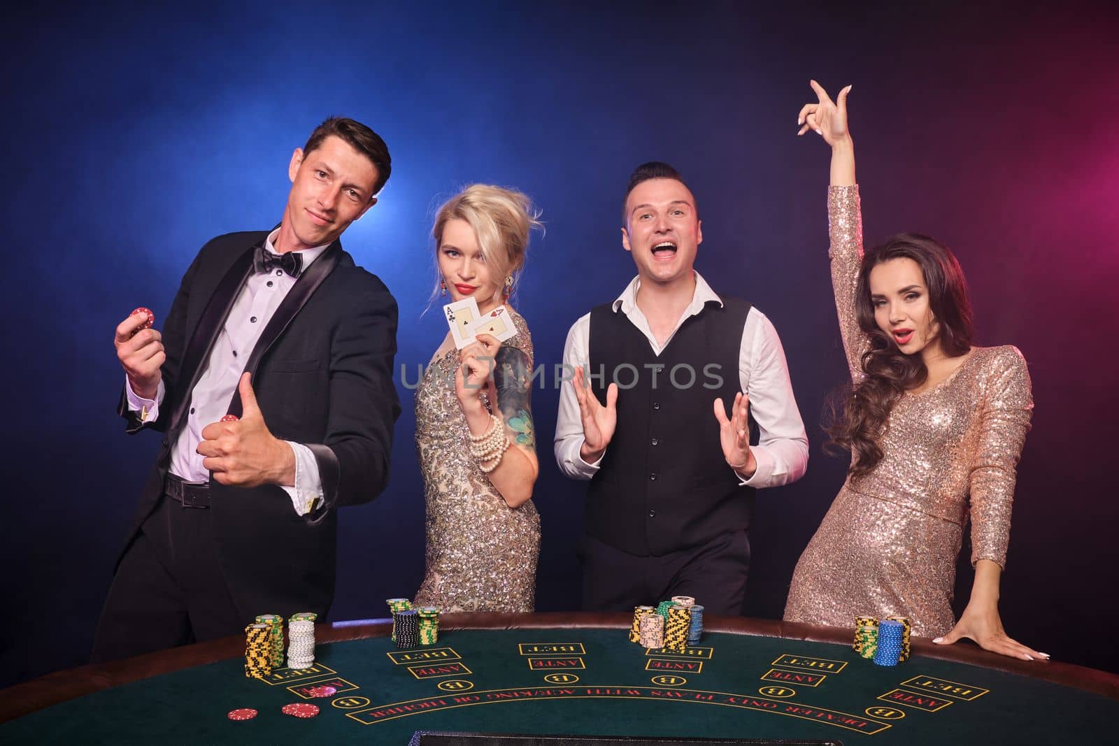 Group of a stylish rich colleagues are playing poker at casino. Youth are making bets waiting for a big win. They are looking excited standing at the table against a red and blue backlights on black background. Risky gambling entertainment.