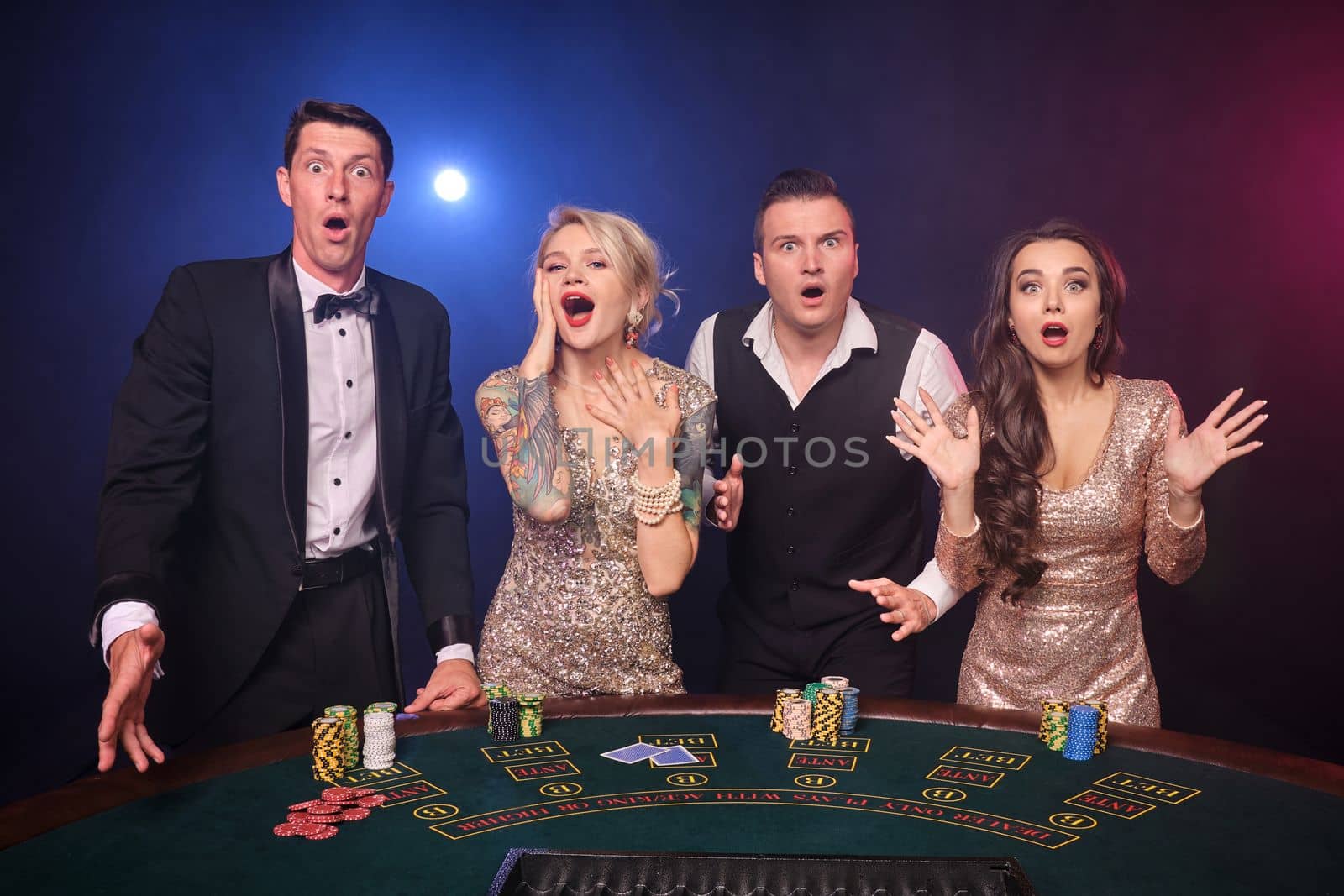 Group of a stylish rich friends are playing poker at casino. Youth are making bets waiting for a big win. They are standing at the table against a red and blue backlights on black background. Risky gambling entertainment.