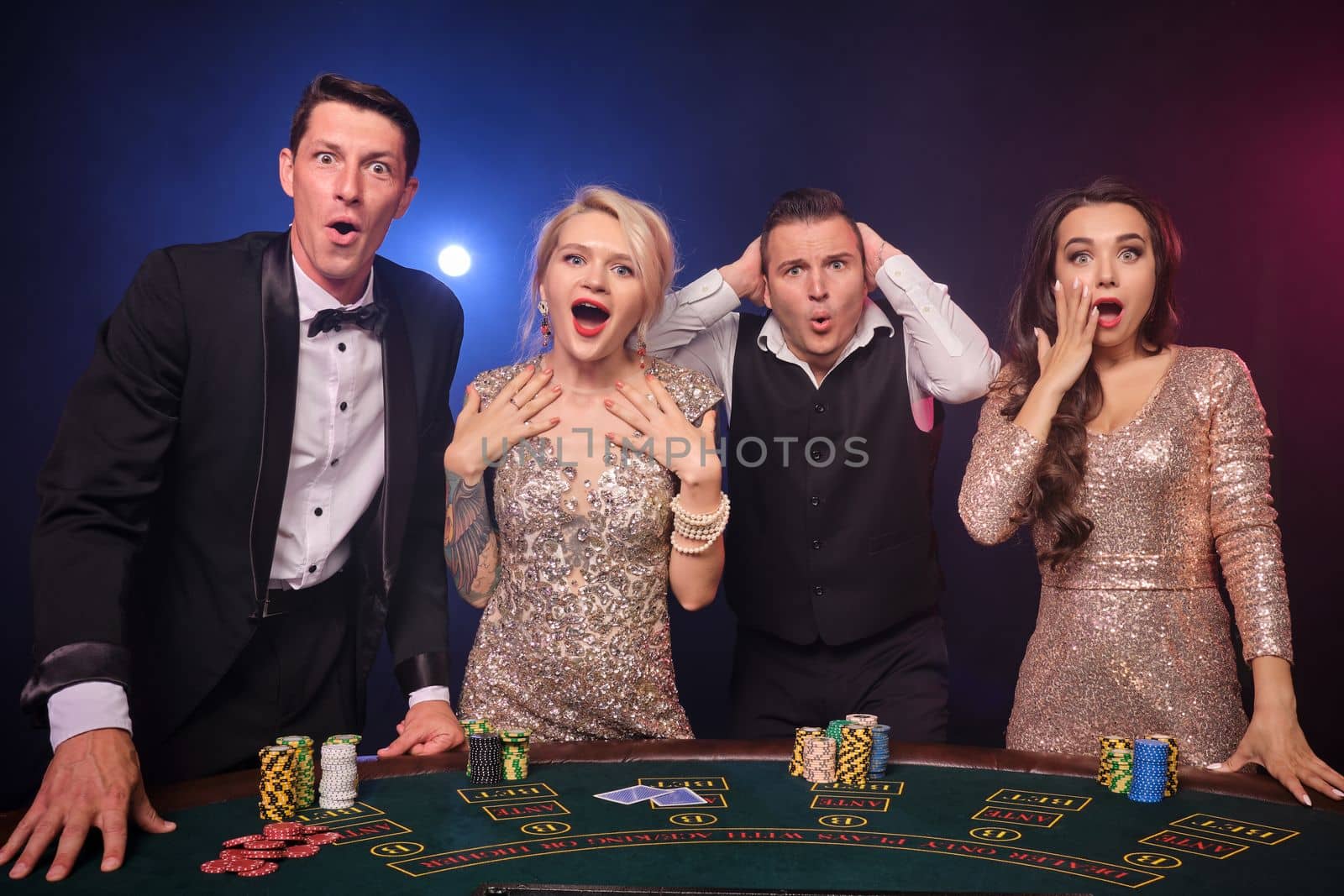 Group of a wondered rich companions are playing poker at casino. Youth are making bets waiting for a big win. They are looking shocked standing at the table against a red and blue backlights on black background. Risky gambling entertainment.