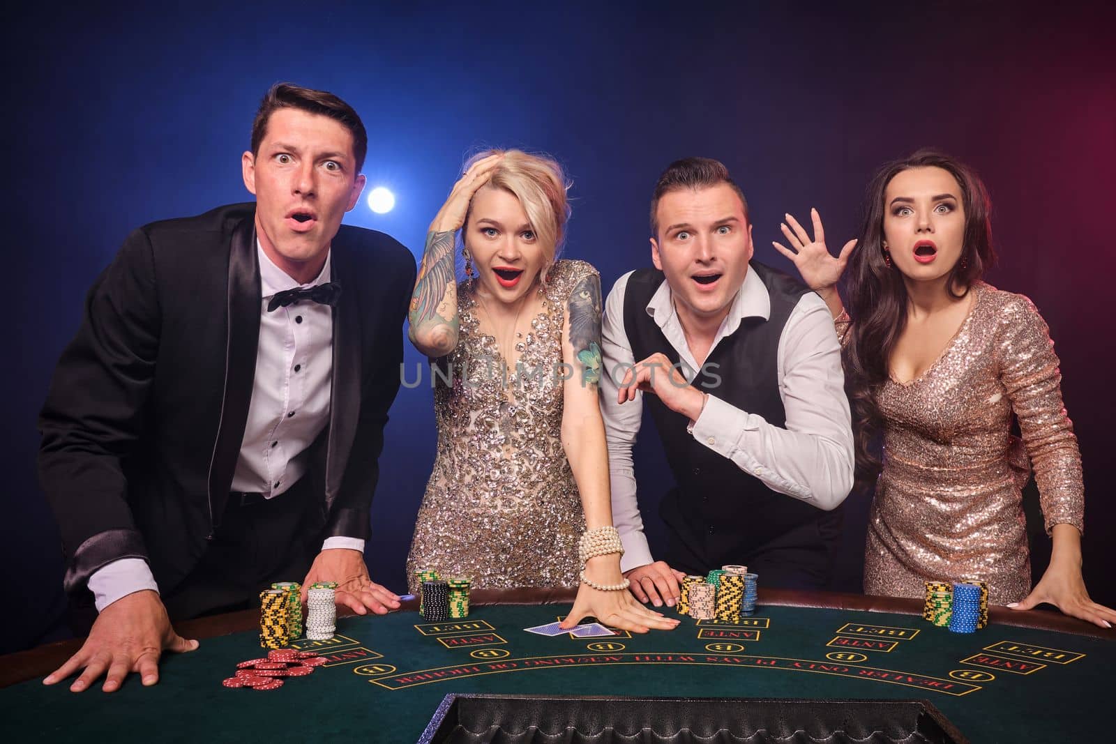 Group of a wondered rich classmates are playing poker at casino. Youth are making bets waiting for a big win. They are looking amazed standing at the table against a red and blue backlights on black background. Risky gambling entertainment.