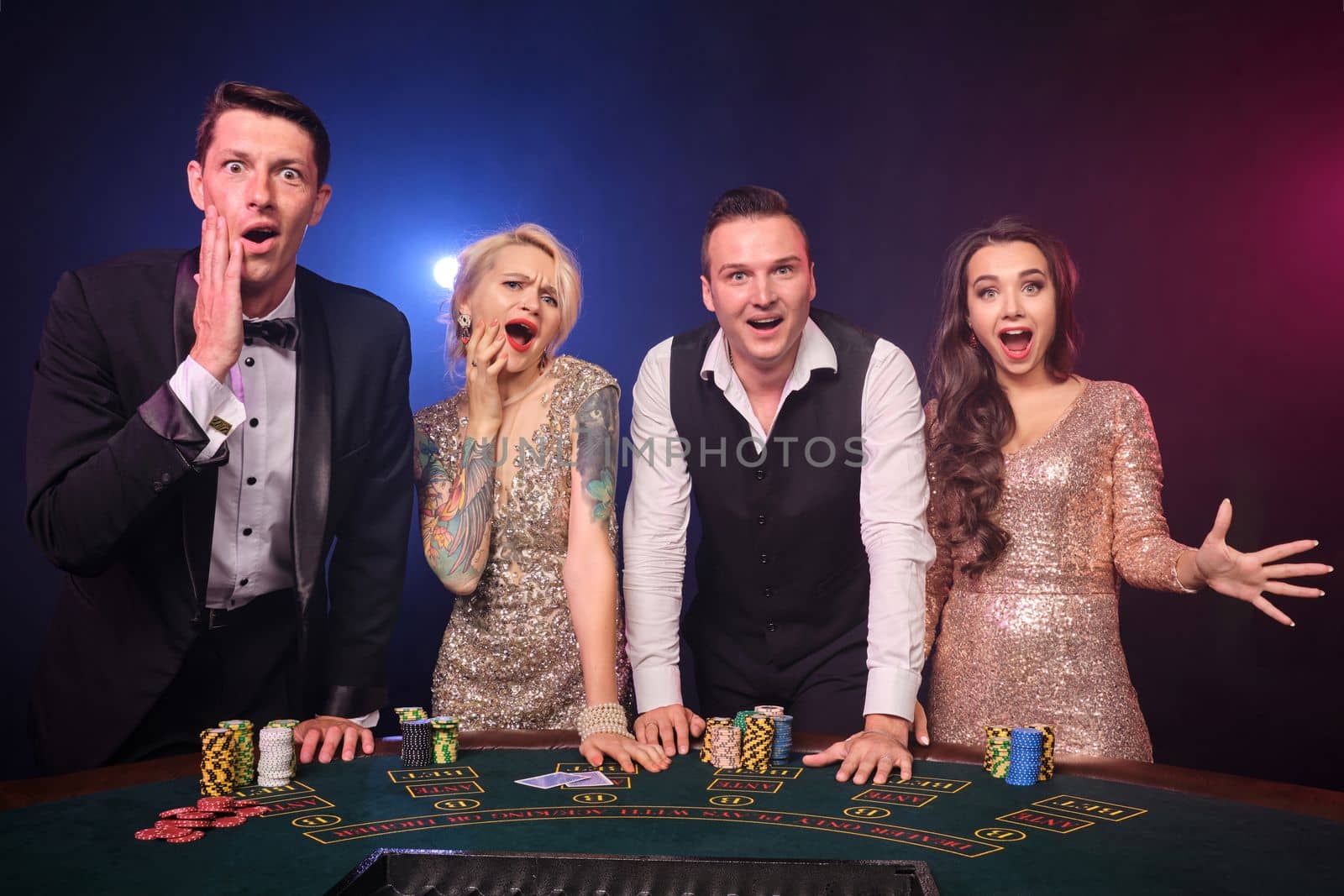 Group of a rich friends are playing poker at casino. Youth are making bets waiting for a big win. They are standing at the table against a red and blue backlights on black background. Risky gambling entertainment.