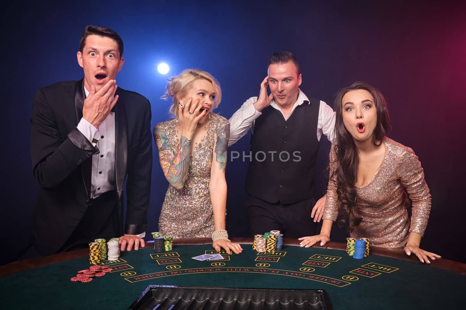 Group of a wondered rich friends are playing poker at casino. Youth are making bets waiting for a big win. They are looking at the camera standing at the table against a red and blue backlights on black background. Risky gambling entertainment.