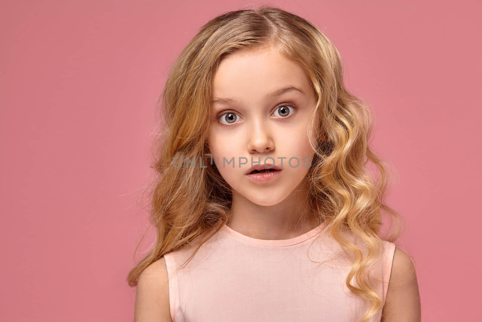 Beautiful little girl with a blond curly hair, in a pink dress is posing for the camera and looks wondered, on a pink background