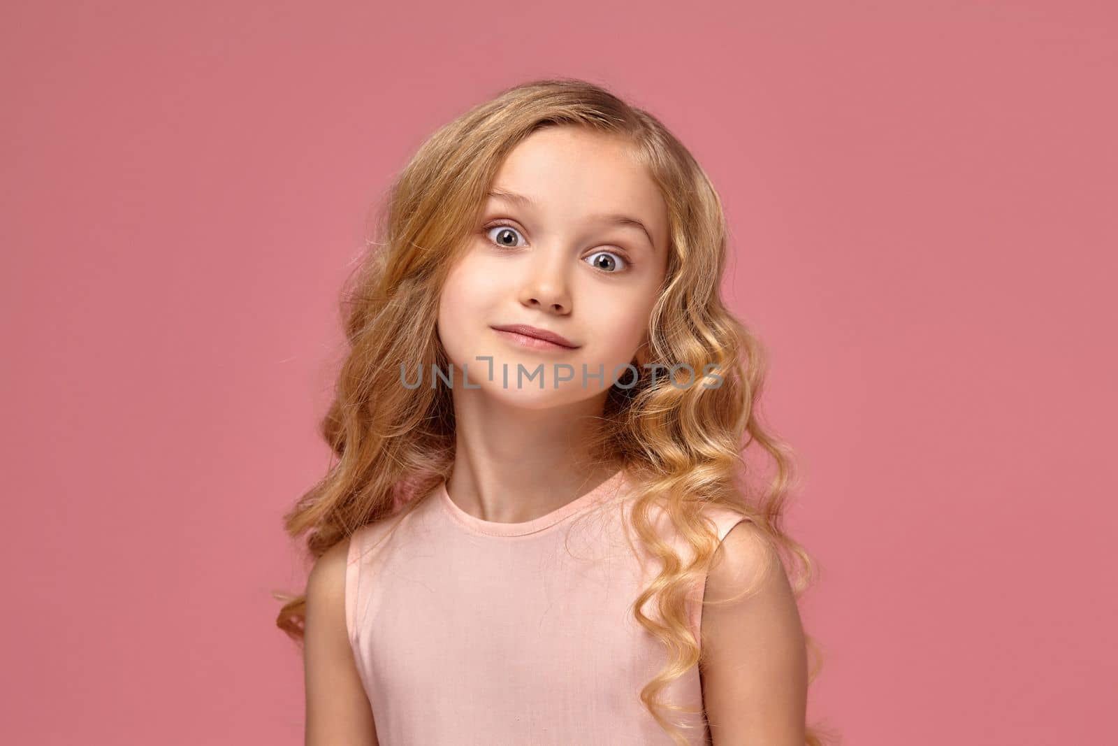 Beautiful little girl with a blond curly hair, in a pink dress poses for the camera and looks shoked, on a pink background