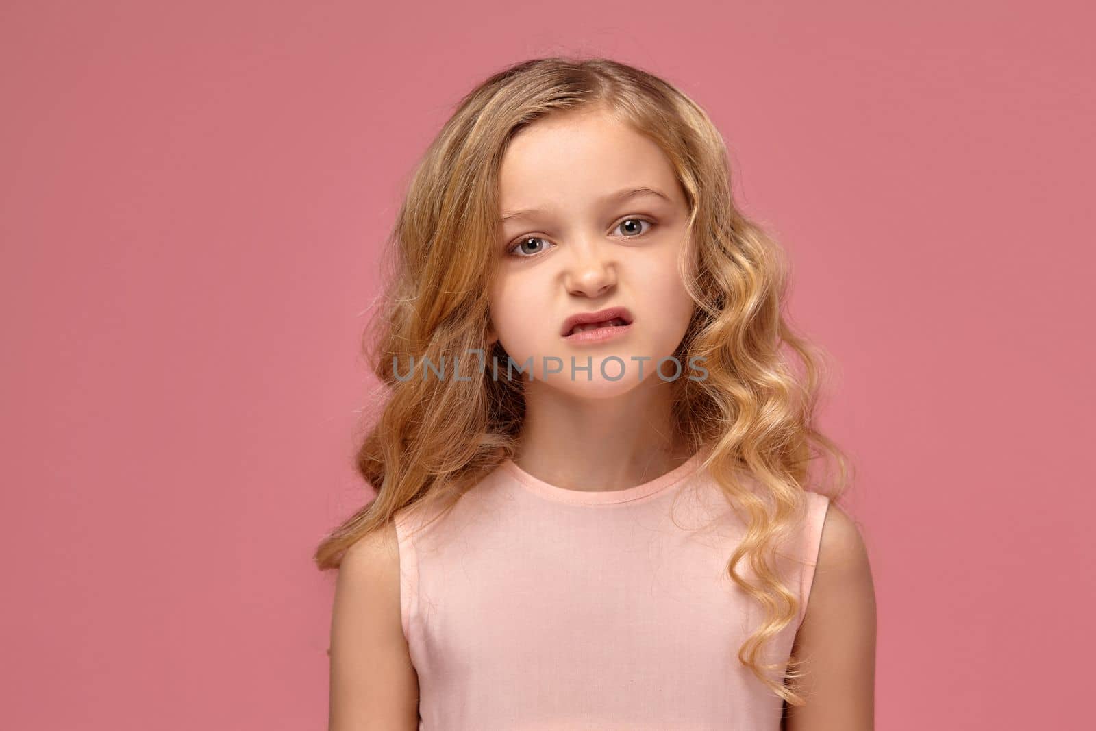 Beautiful little girl with a blond curly hair, in a pink dress poses for the camera and looks angry, on a pink background