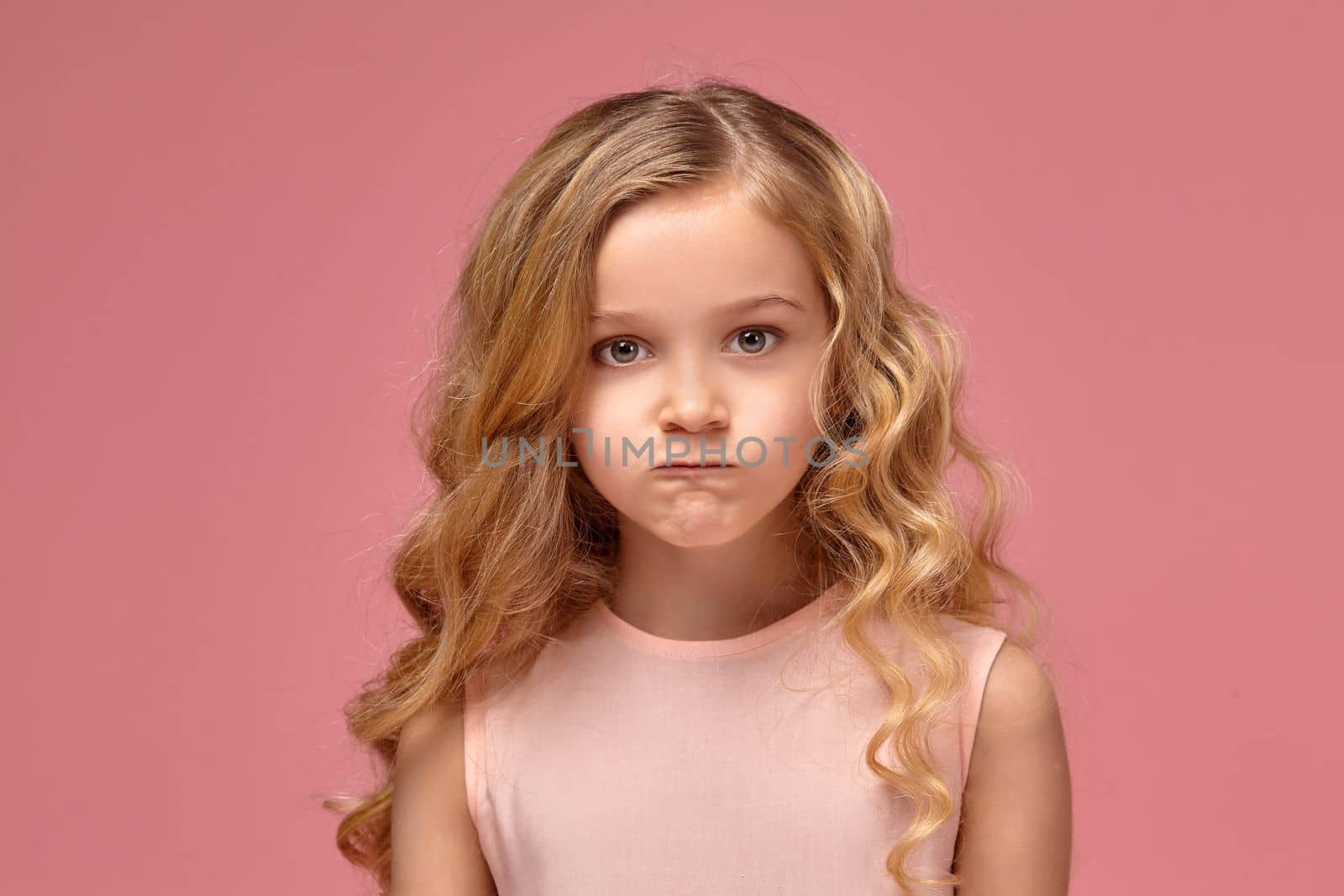 Pretty little girl with a blond curly hair, in a pink dress is posing for the camera and looking angry, on a pink background